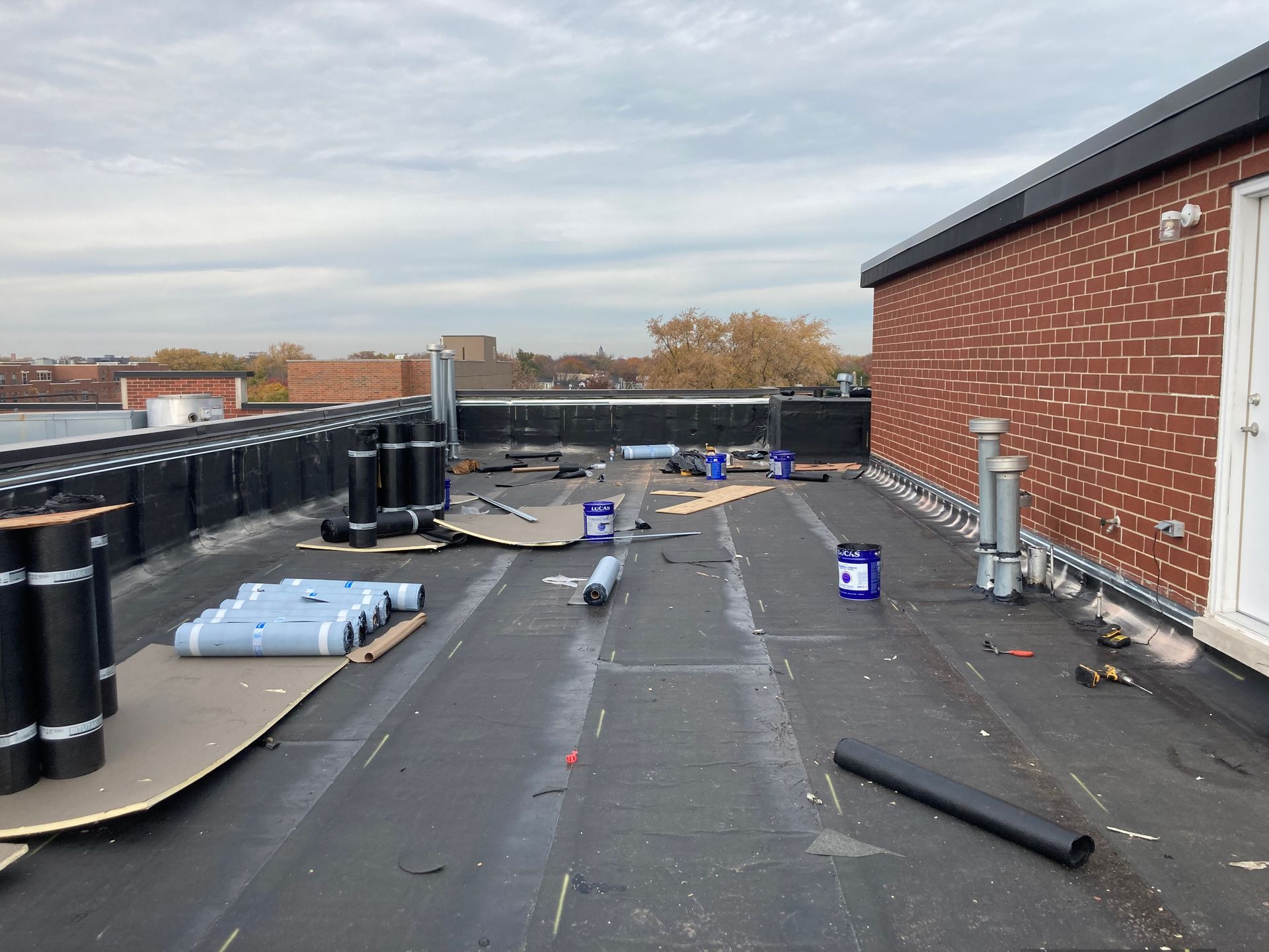 Flat commercial roof under construction; rolls of black roofing material and tools visible.