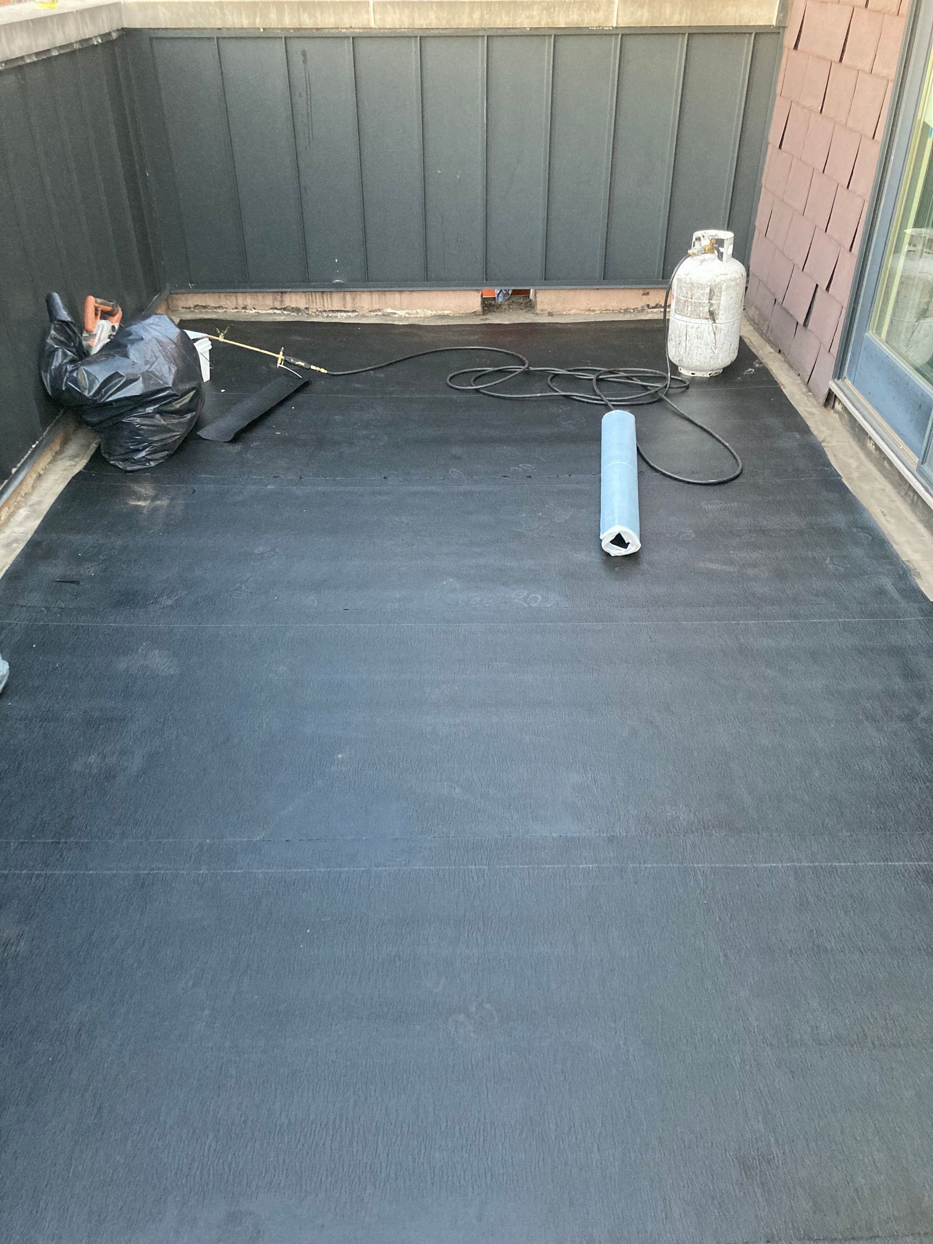Black rubber-covered deck with propane tank, trash bag, and blue object. Dark gray wooden walls.