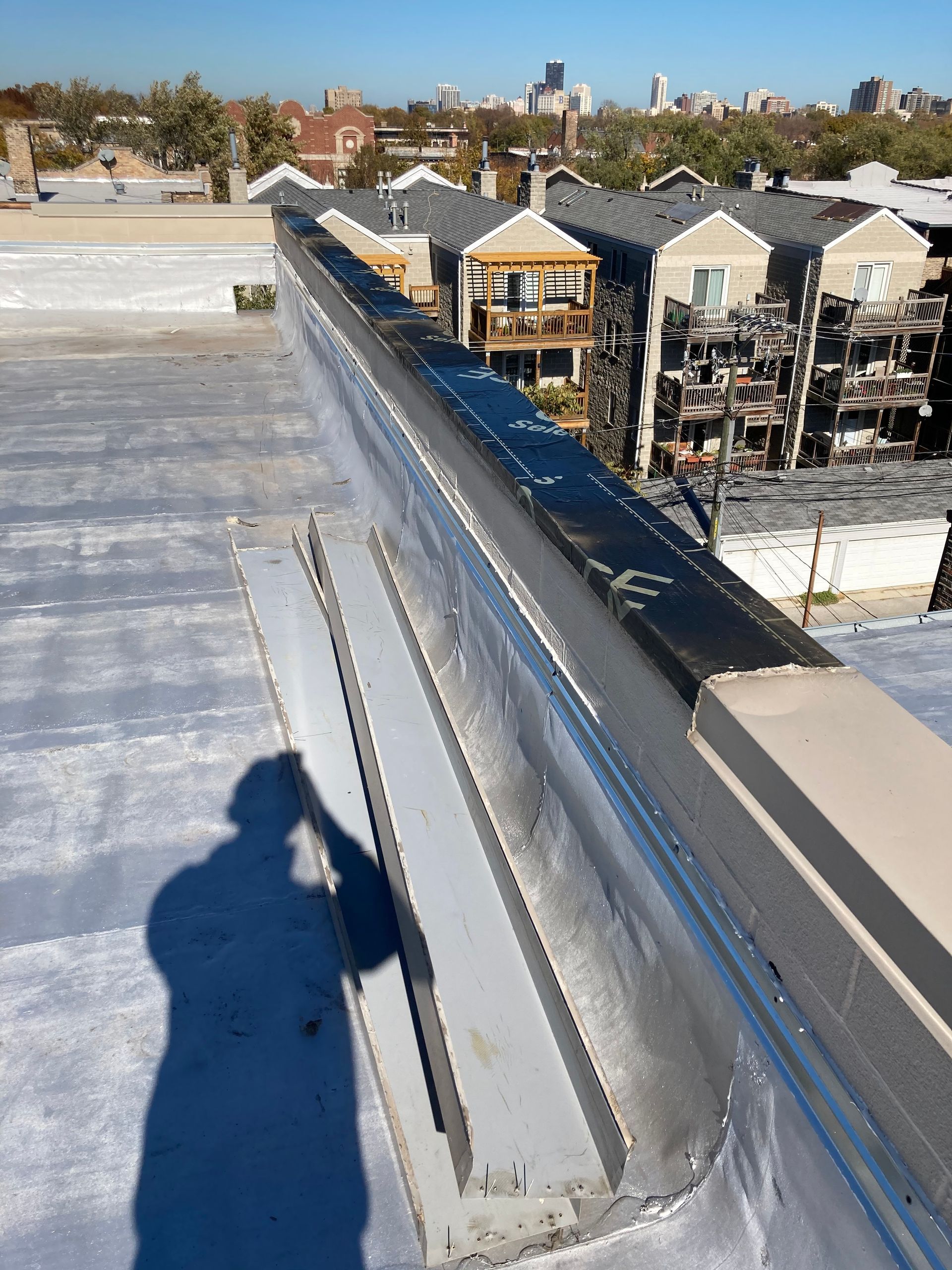 A person's shadow on a rooftop, with a metal flashing edge, and cityscape in the background.