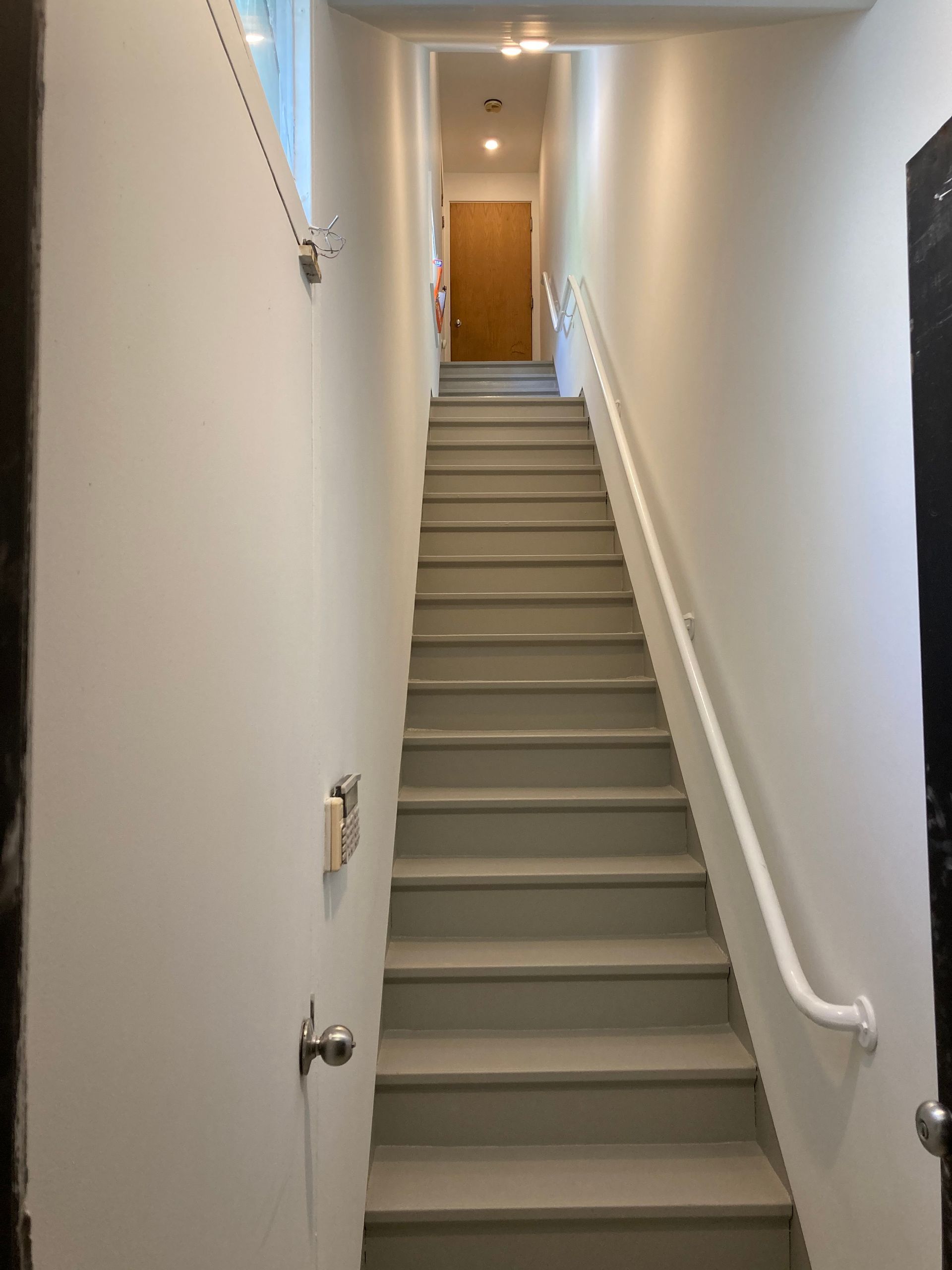 Narrow staircase with grey treads and white walls, leading to a closed wooden door. Handrail on the right.