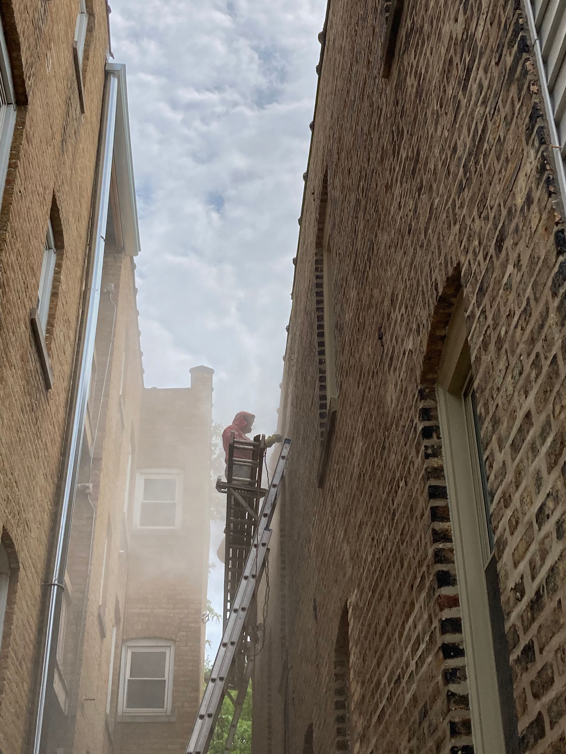 Firefighter on a ladder between brick buildings, smoke visible, cloudy sky.