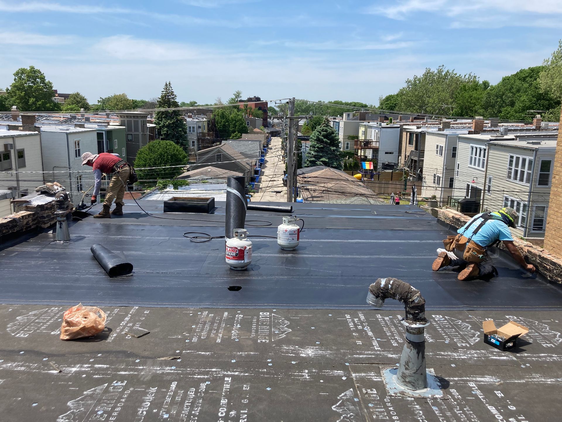 Construction workers on a flat roof, installing black roofing material on a sunny day.