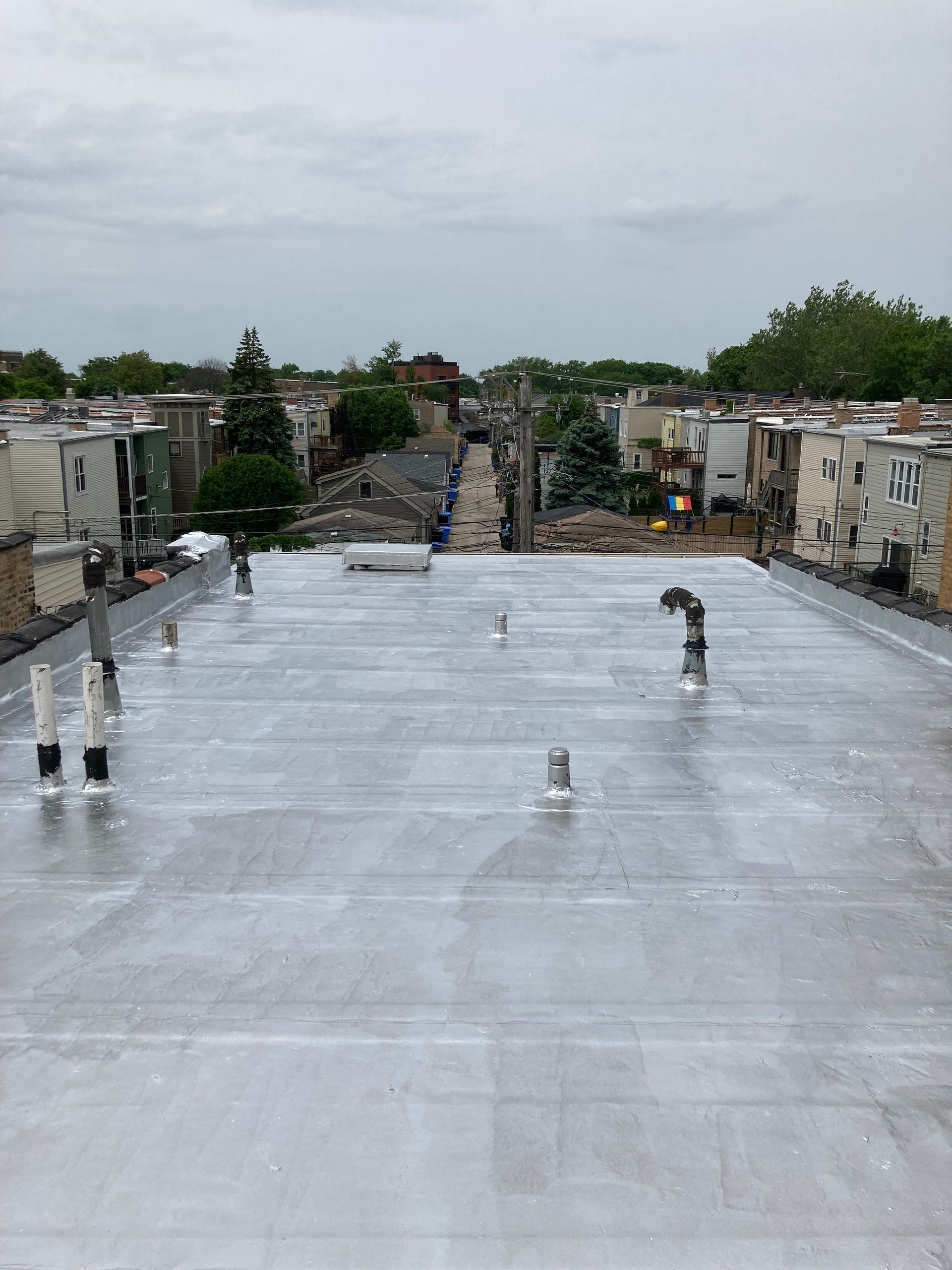 View of a flat, gray rooftop with plumbing vents, overlooking a residential street under a cloudy sky.
