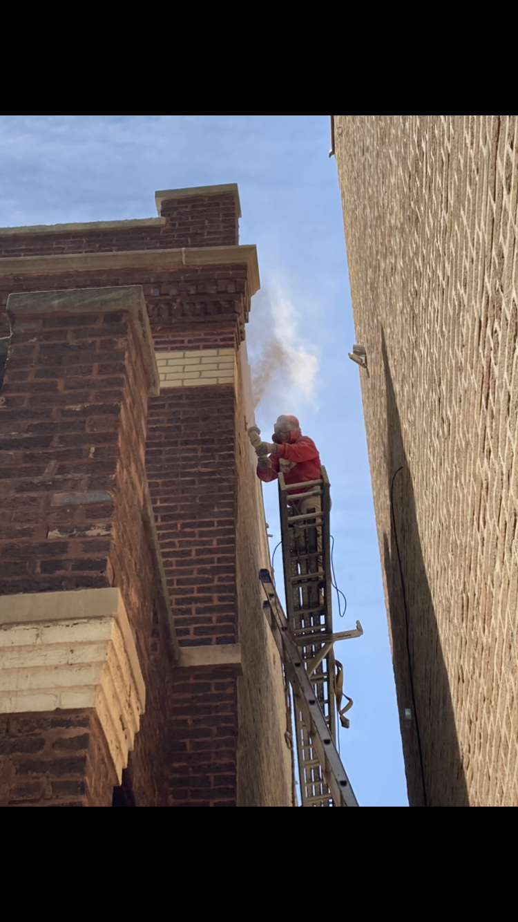 Firefighter on a ladder sprays water at a building. Smoke rises. Blue sky in background.