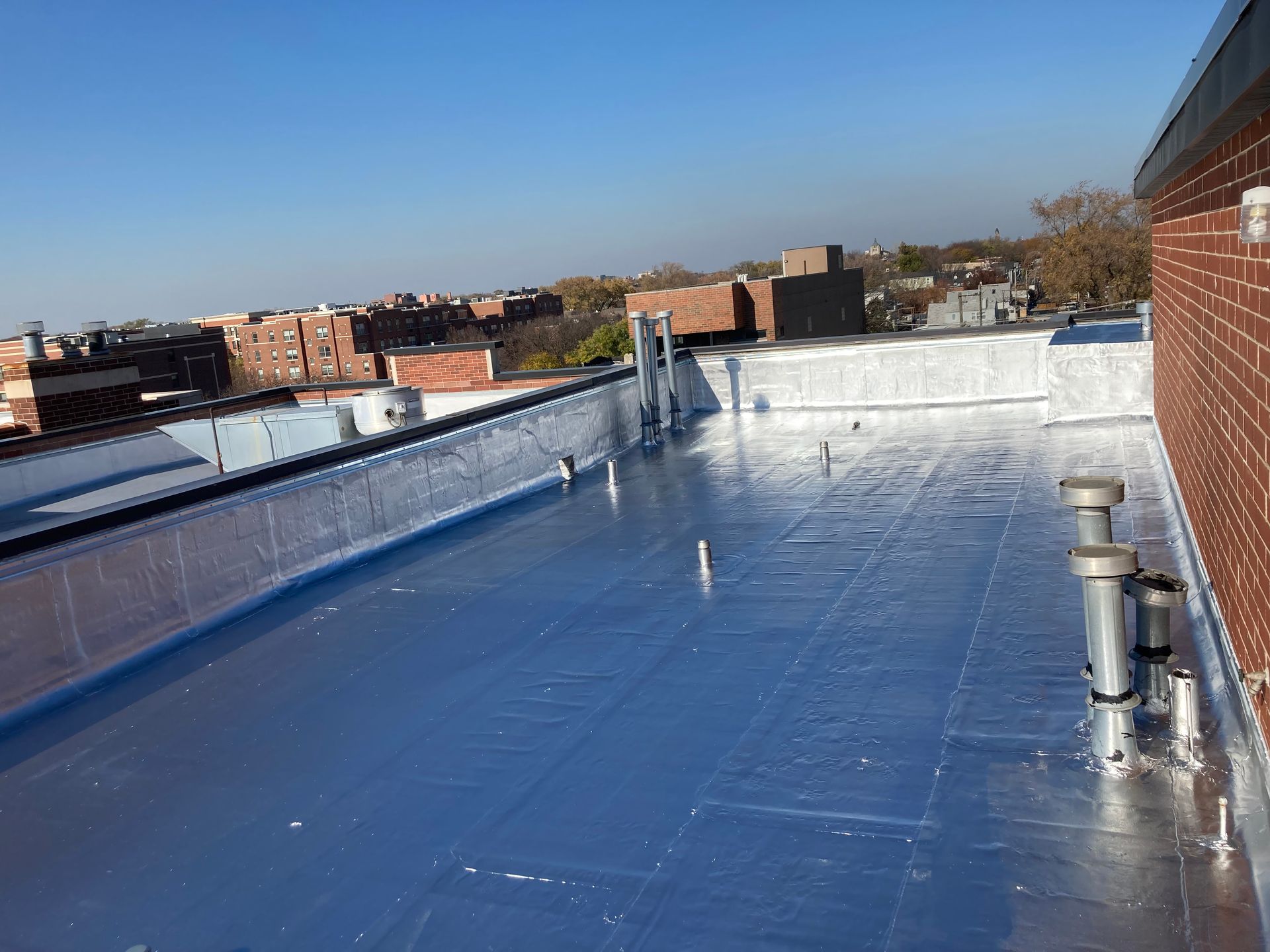 Flat, shiny blue roof with silver edging, near brick building and cityscape on a clear day.
