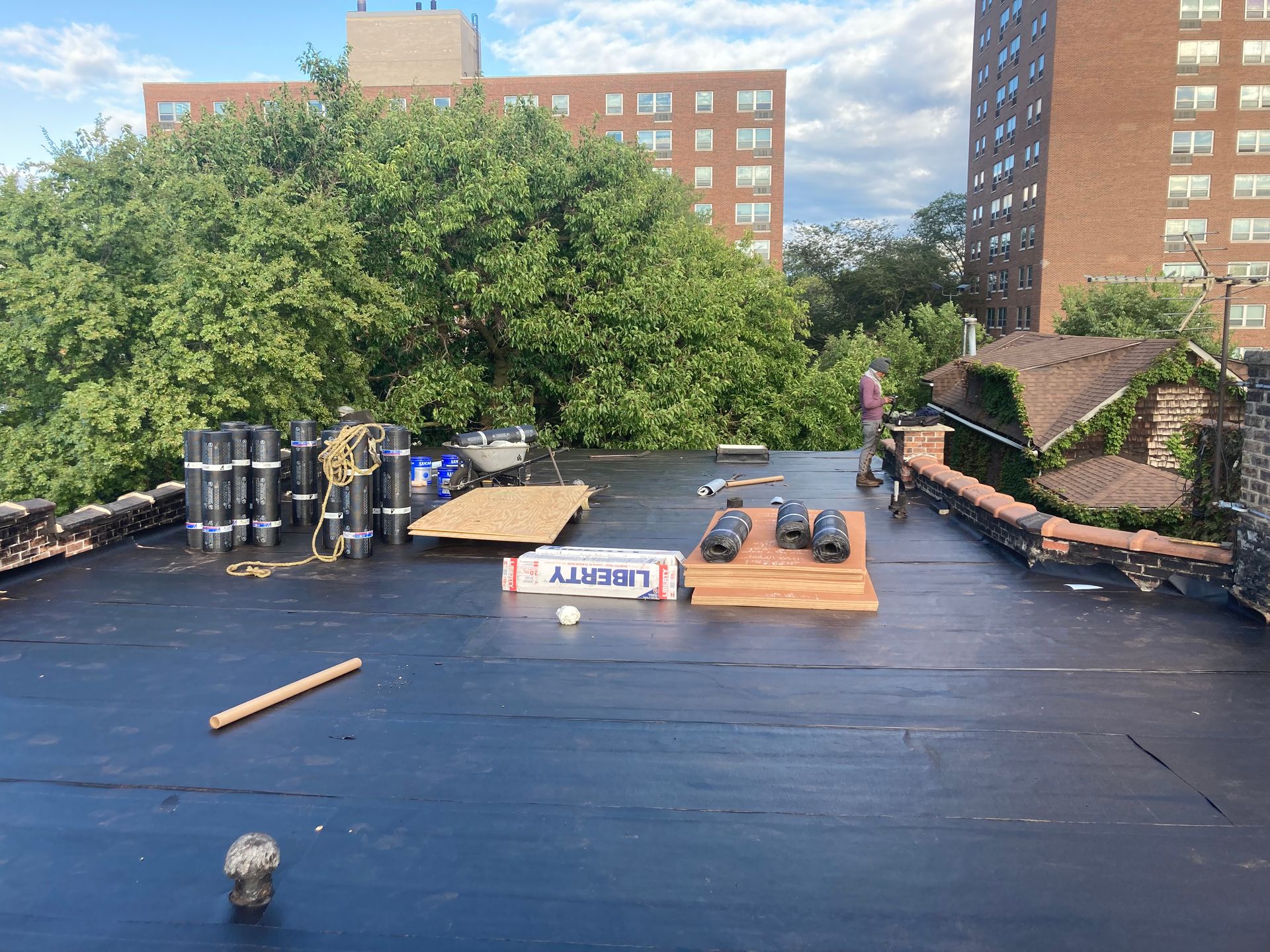 Rooftop with roofing materials: rolls, boards, Liberty label, and tools, with buildings and trees in the background.
