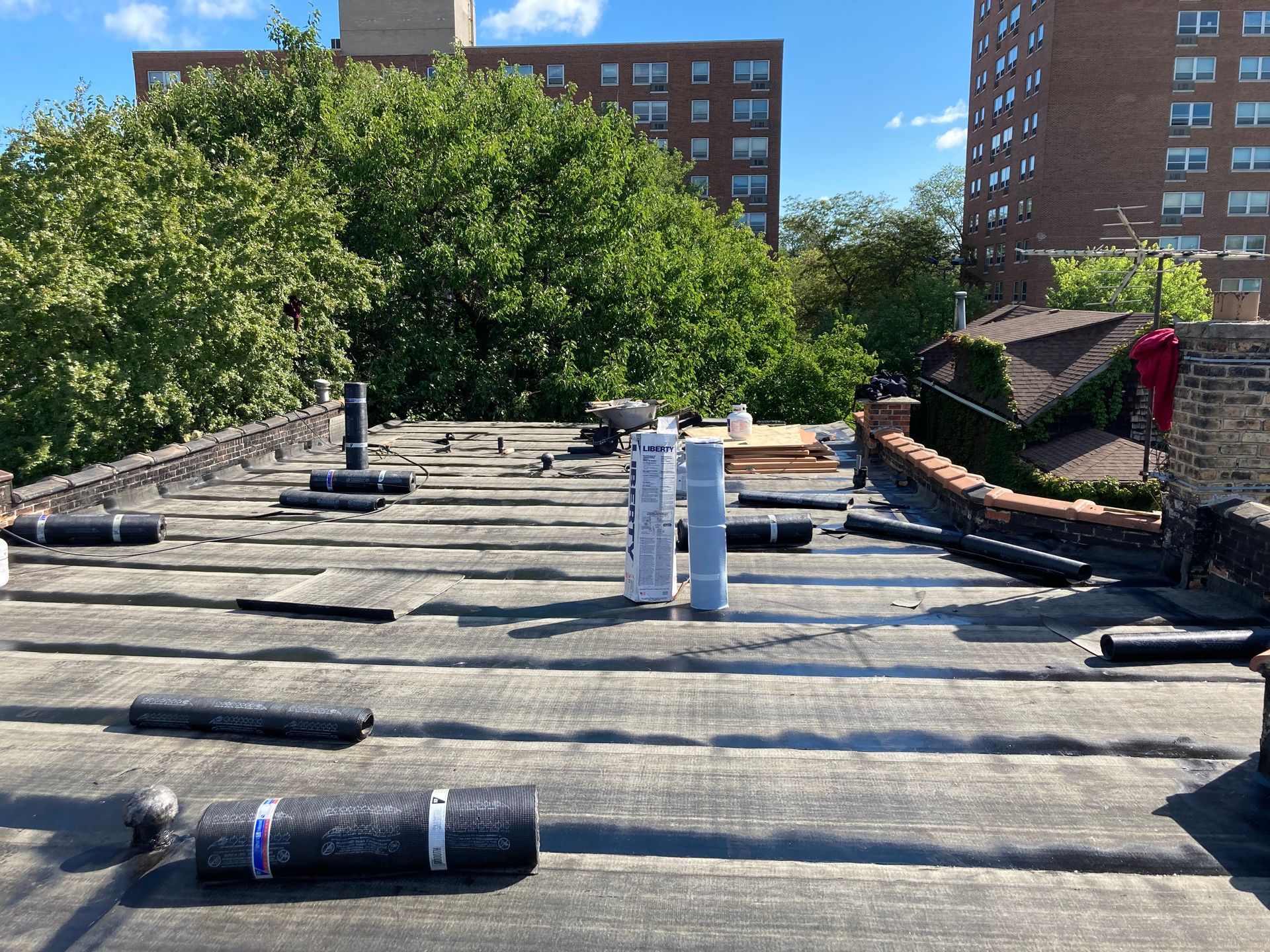 Flat rooftop with pipes and rolled roofing material; apartment buildings and trees in background.