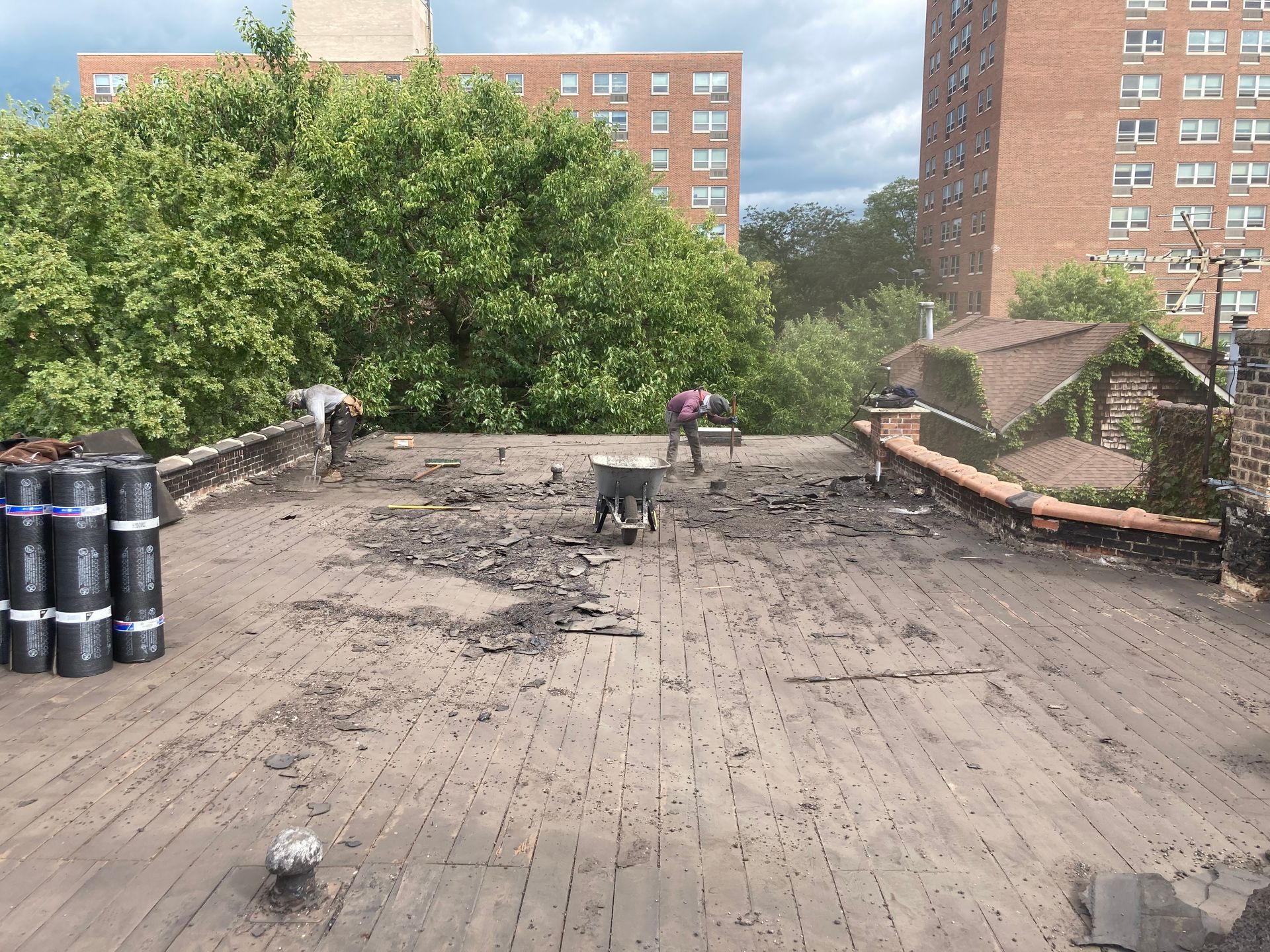 Roofers removing old roofing materials on a city building's flat roof, with stacked rolls and buildings in the background.