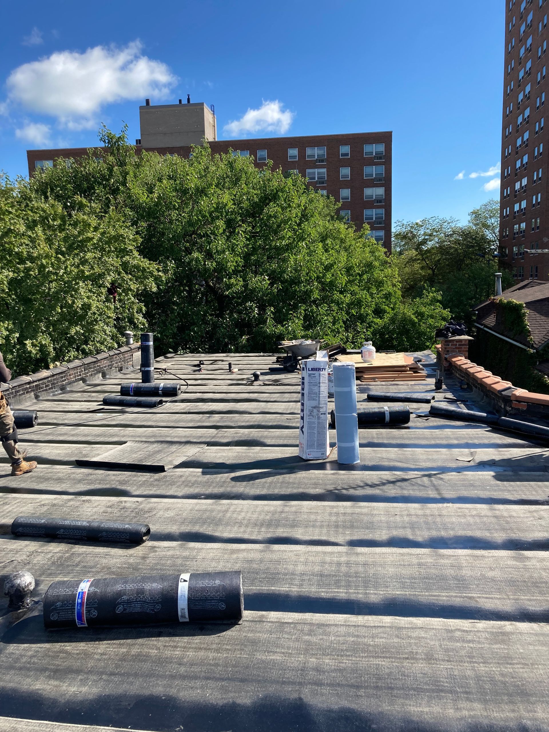 Rooftop with black roofing material, ventilation pipes, and rolls of material. City buildings and trees in the background.