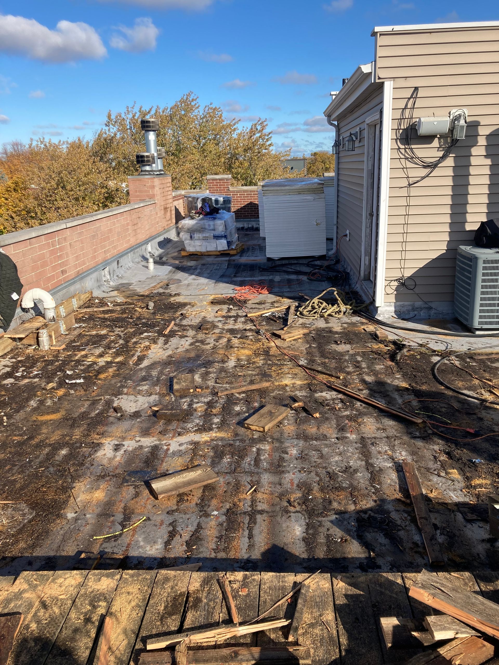 Rooftop with damaged wood surface, materials, and a brick chimney under a blue sky.