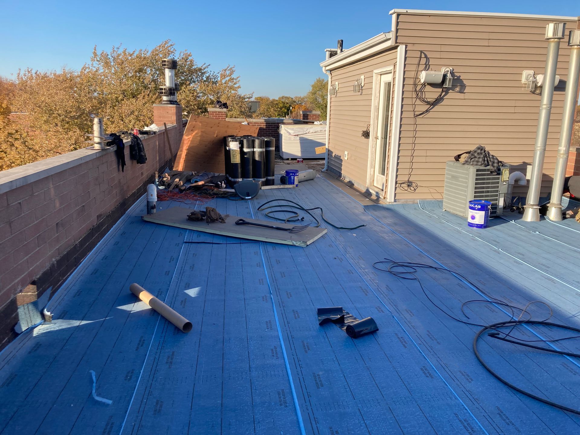 Rooftop with blue waterproofing, building access door, brick chimney, and construction materials under clear sky.