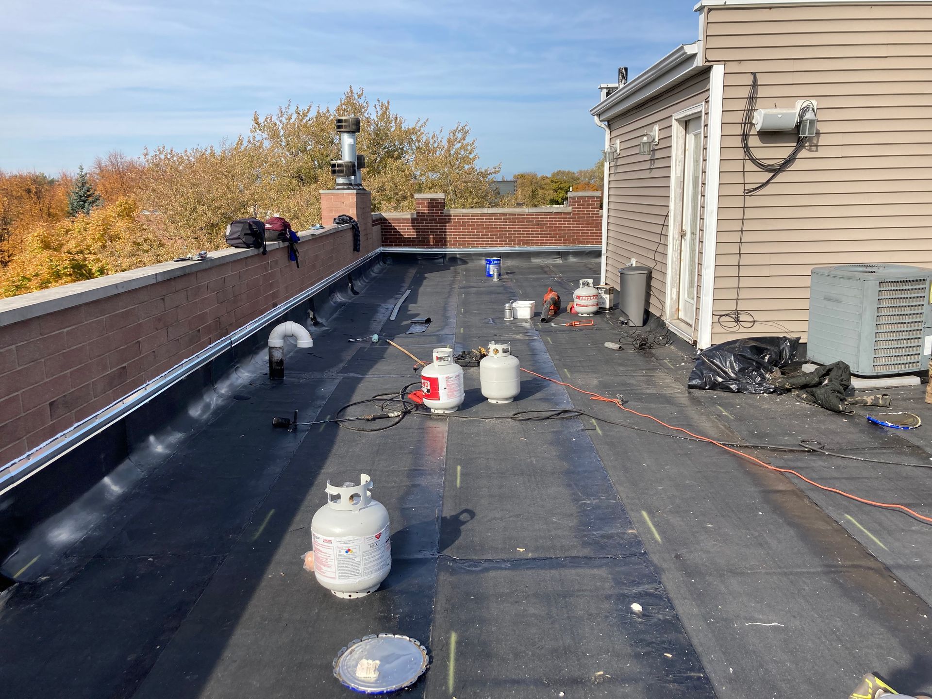Rooftop with roofing supplies and propane tanks; brick wall, trees, and building in the background.