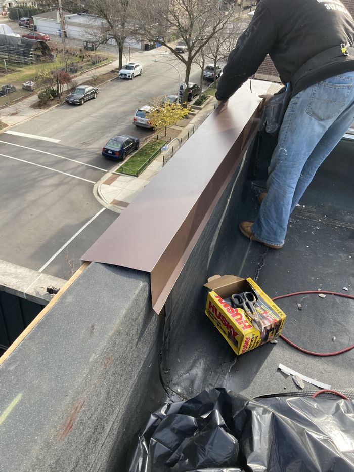 Roofer installing brown metal flashing on a flat roof edge. Tools and street view are visible.