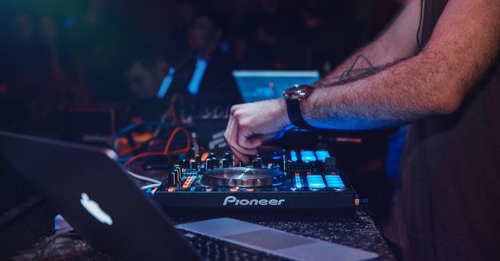 DJ at a club, mixing music on a Pioneer controller, hands on knobs, laptop in the foreground.