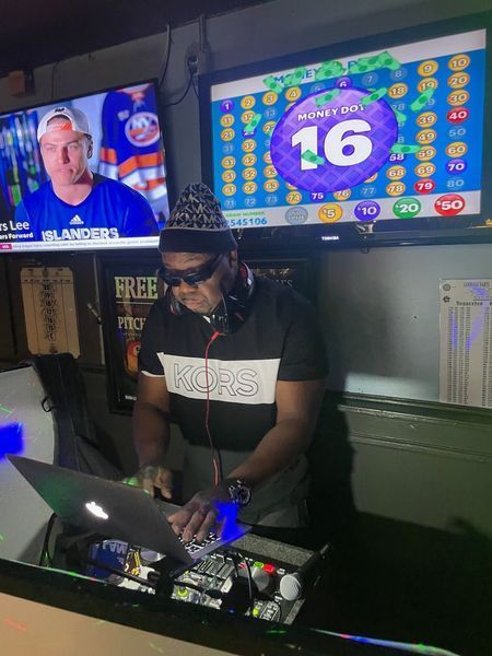 DJ at a bar, wearing headphones, playing music on a laptop, with TVs showing sports and a game.