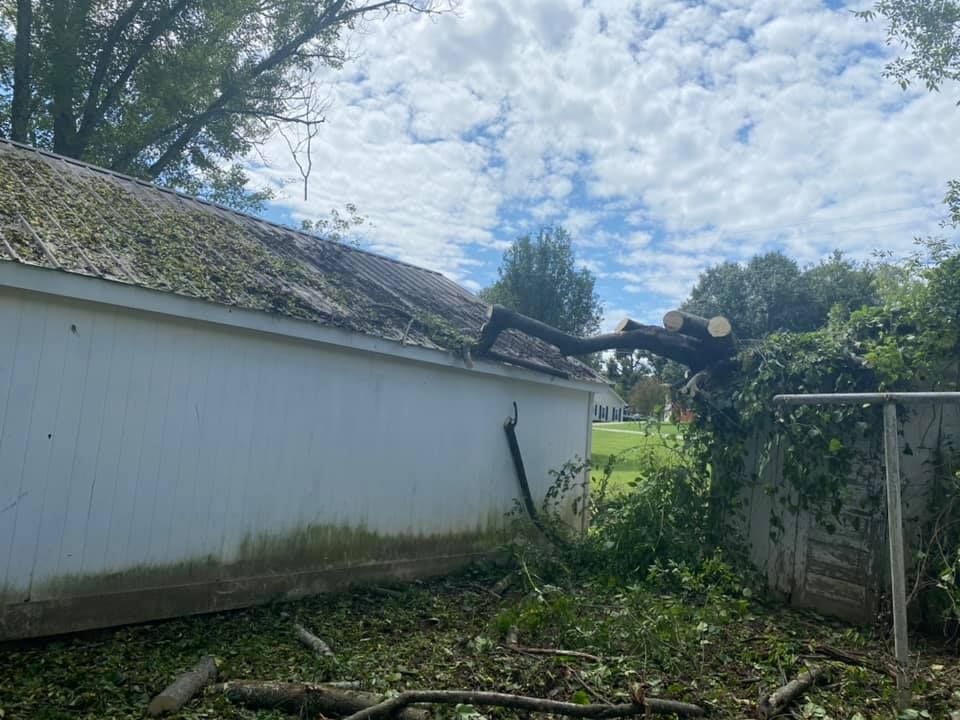 A tree branch rests on a white building's roof, debris scattered below a cloudy sky.