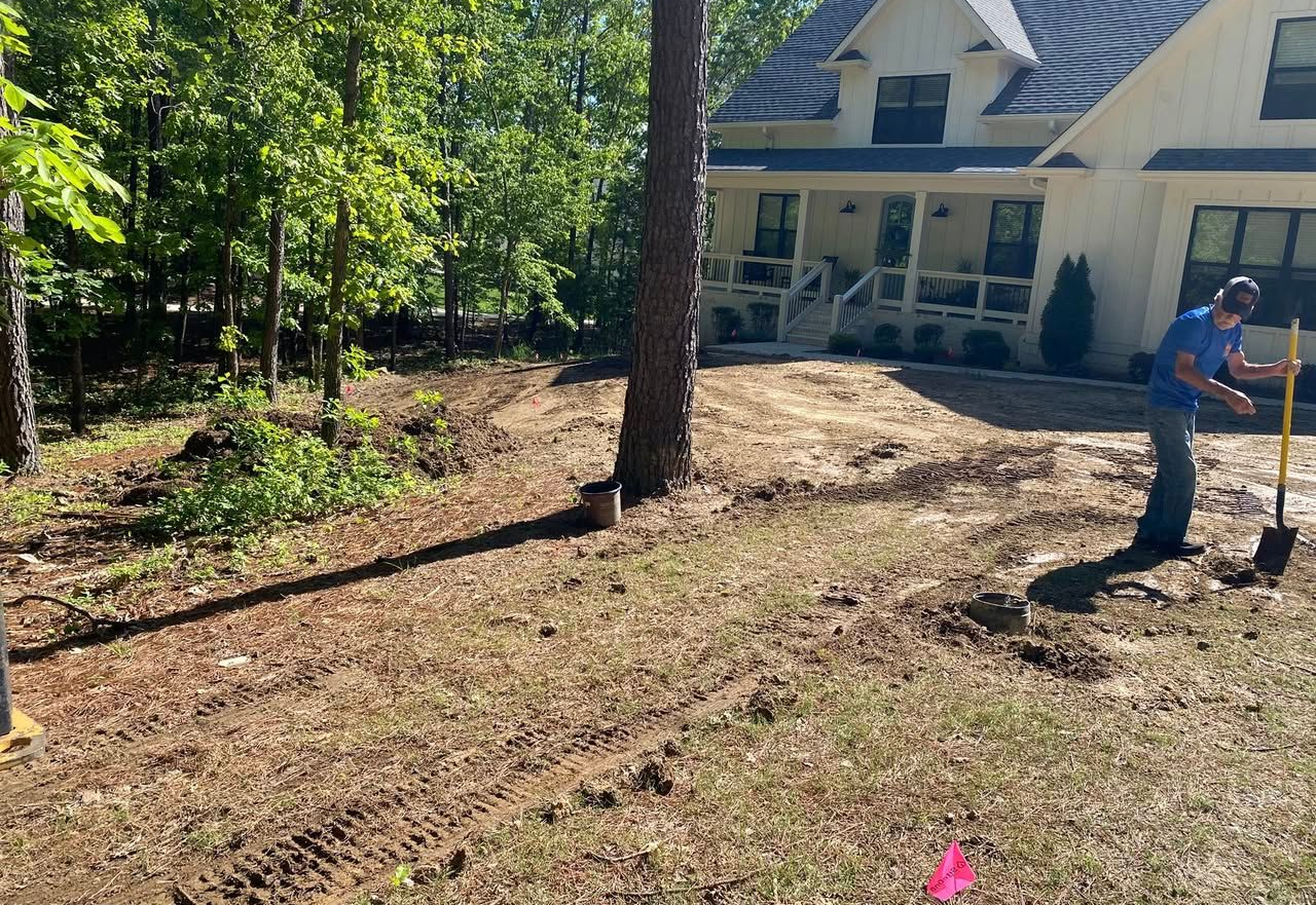 Man shoveling dirt near a house with a porch, trees, and brown soil.