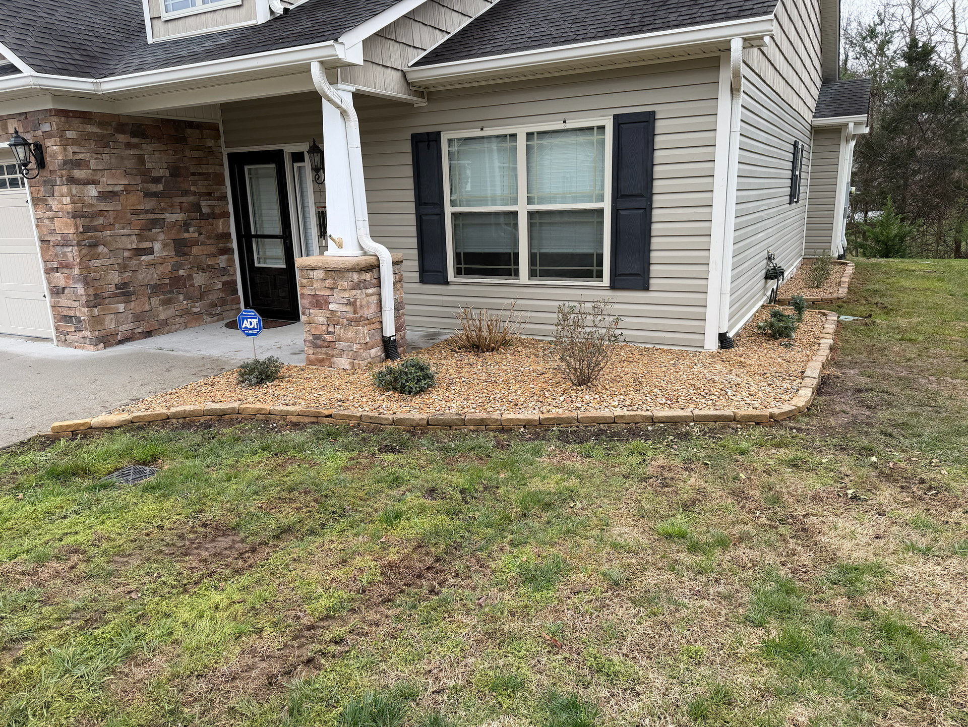 A beige house exterior with stone accents, a covered front porch, and a gravel garden bed in front of a green lawn.