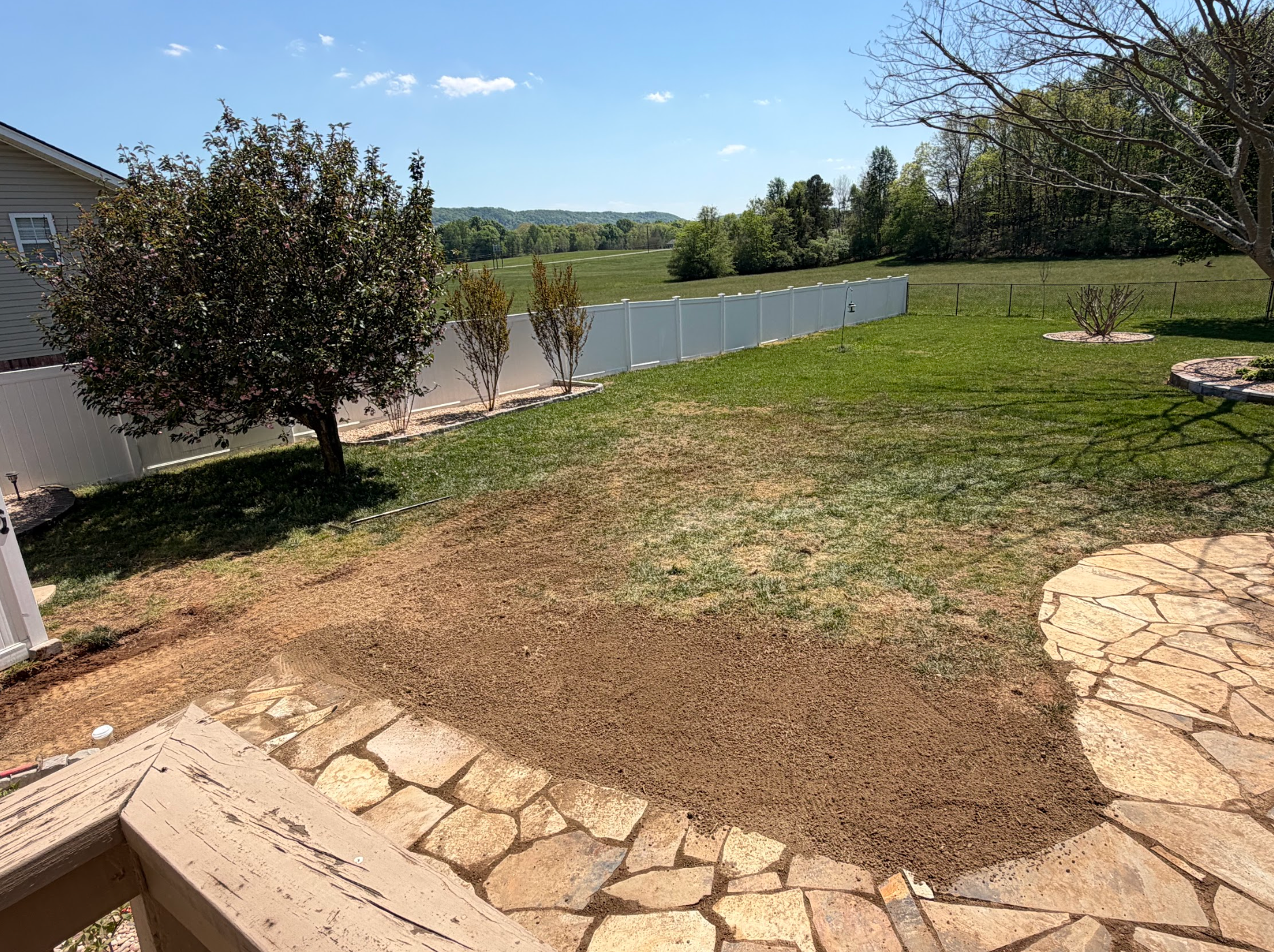 Backyard with patchy grass, stone patio, trees, and a lake or pond view beyond a white fence