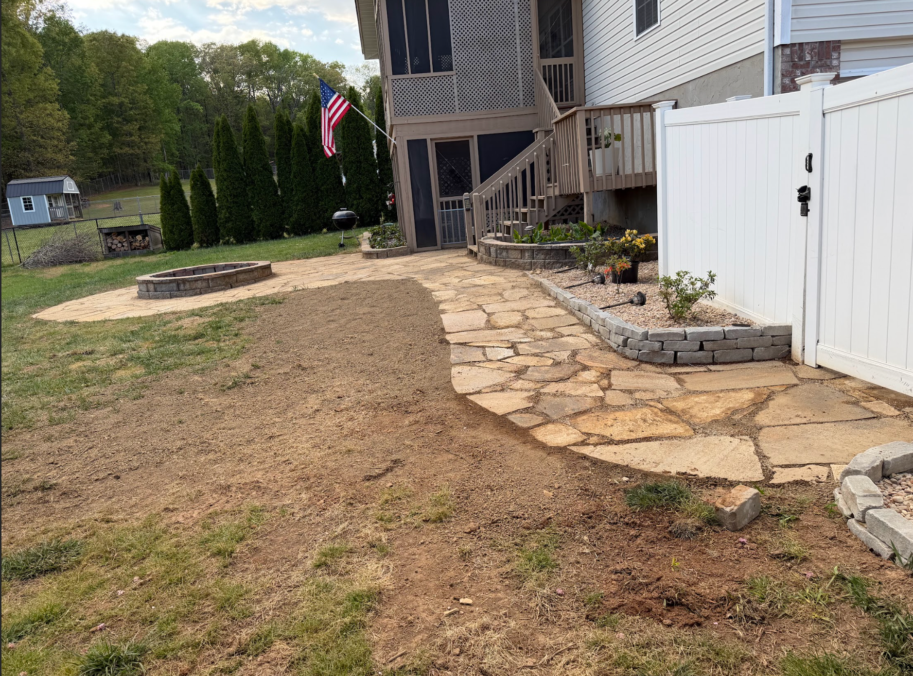 Stone walkway beside a white fence leading to a house, with a flag and grassy yard.