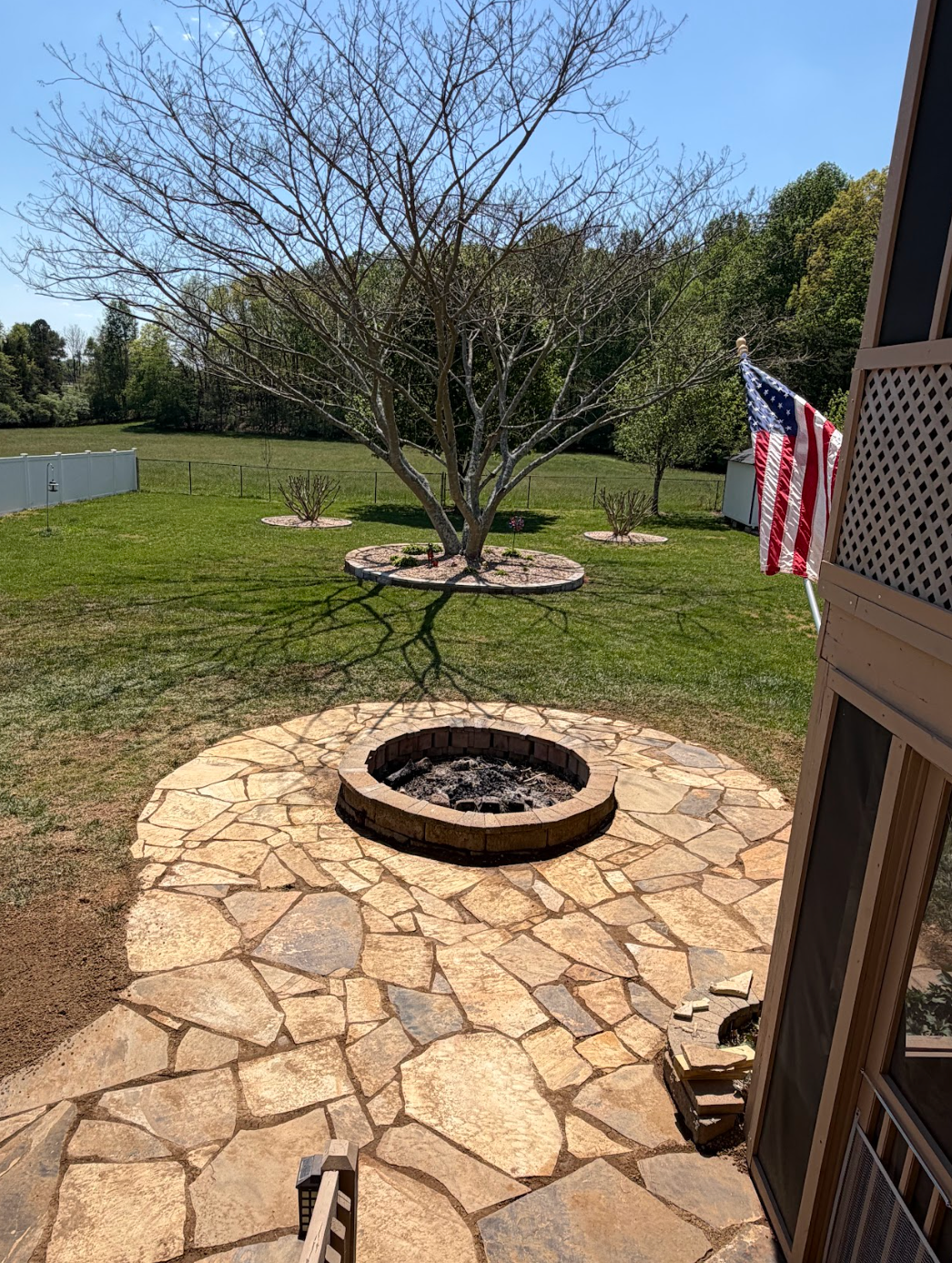 Backyard stone patio with a circular fire pit, leafless tree, and an American flag near the house