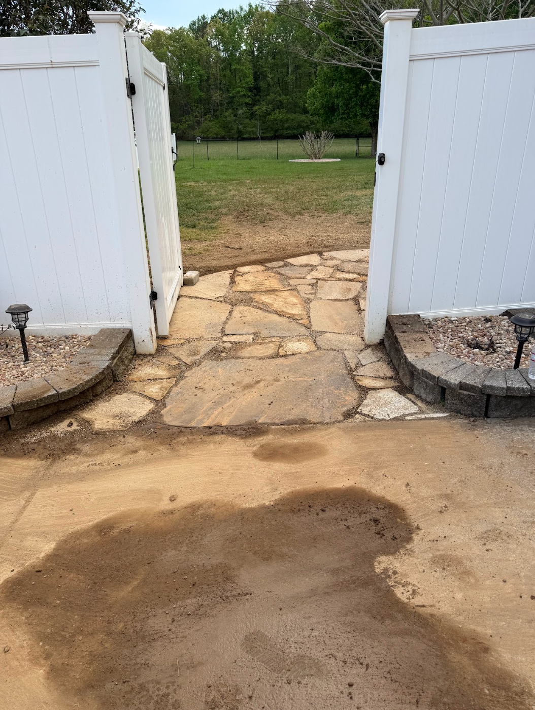 Open white gates framing a stone path to a grassy backyard, with wet concrete in the foreground