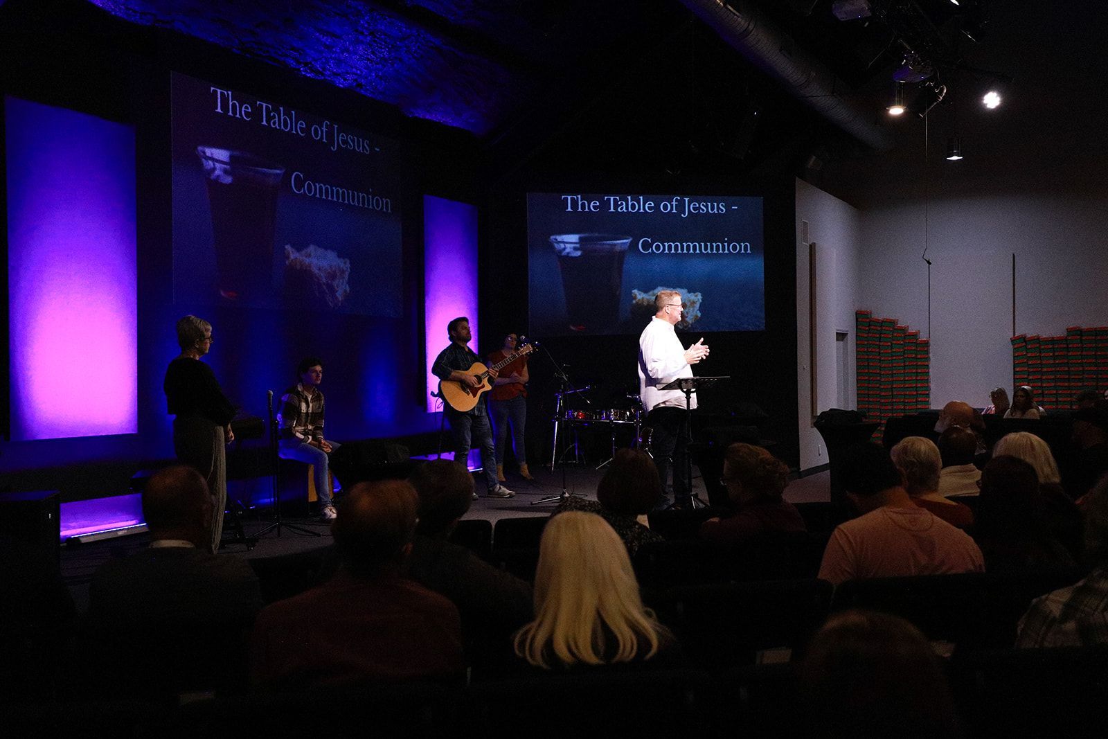 A group of people are sitting in front of a stage in a dark room.