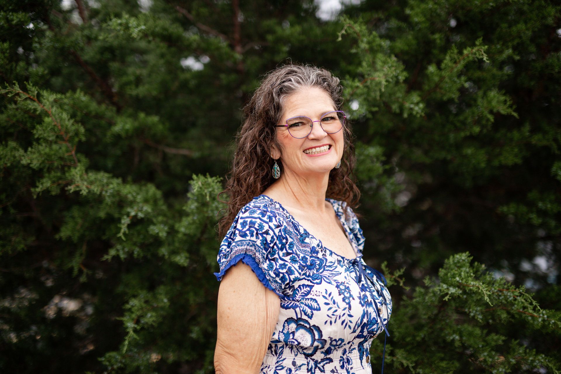 Smiling woman in a blue-and-white blouse standing outdoors in front of green foliage