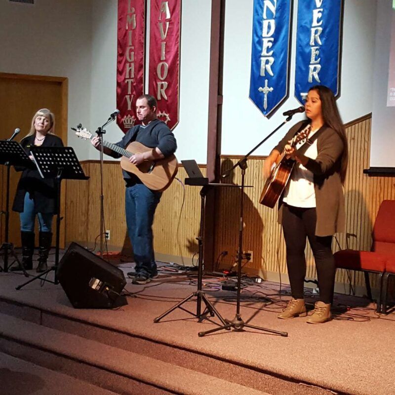 A man playing a guitar and a woman singing in front of a banner that says mighty