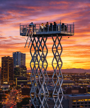 People on a high platform overlook a city skyline at sunset with orange and purple clouds.