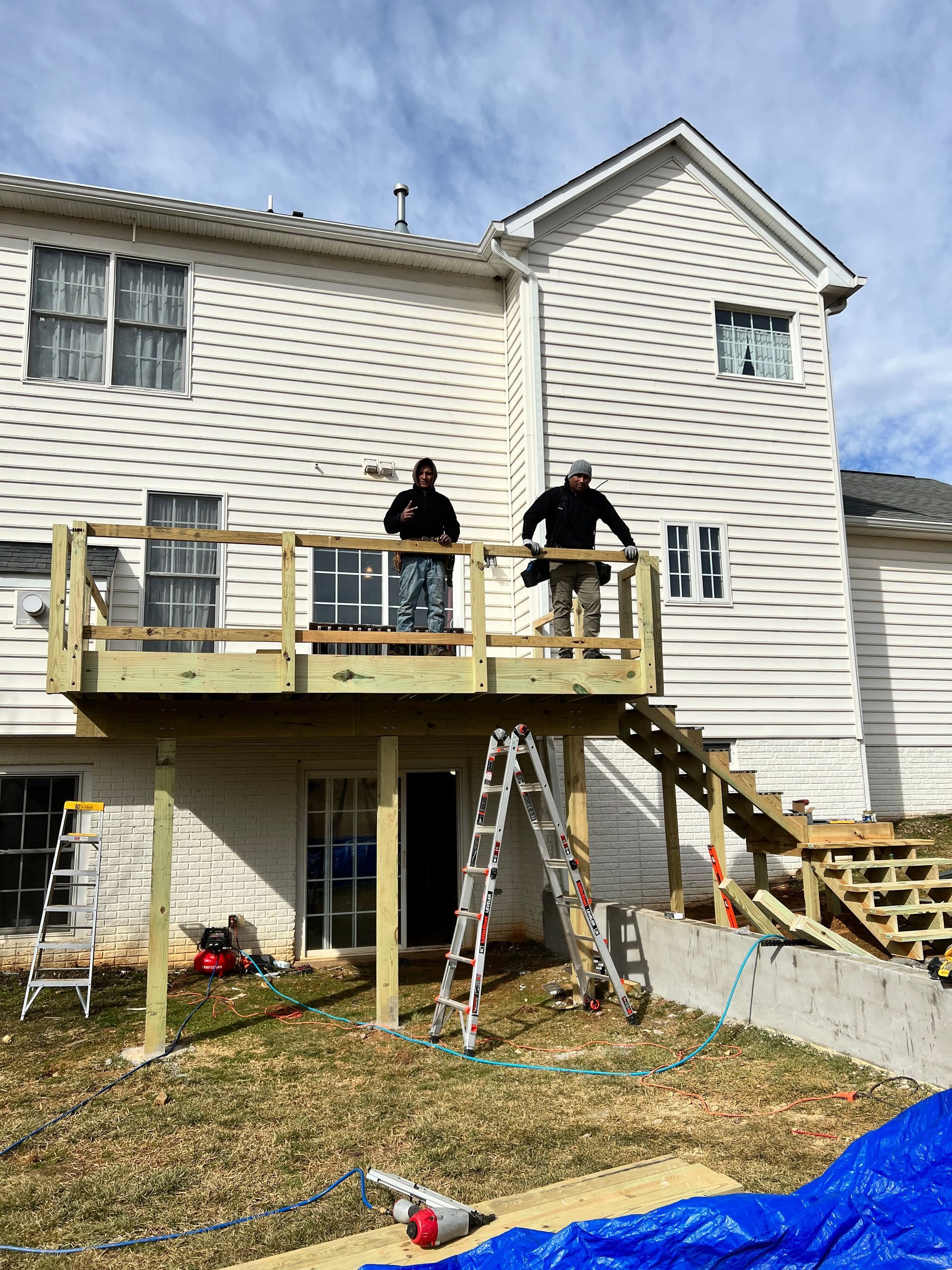 Two workers stand on a newly constructed wooden deck attached to a two-story house.