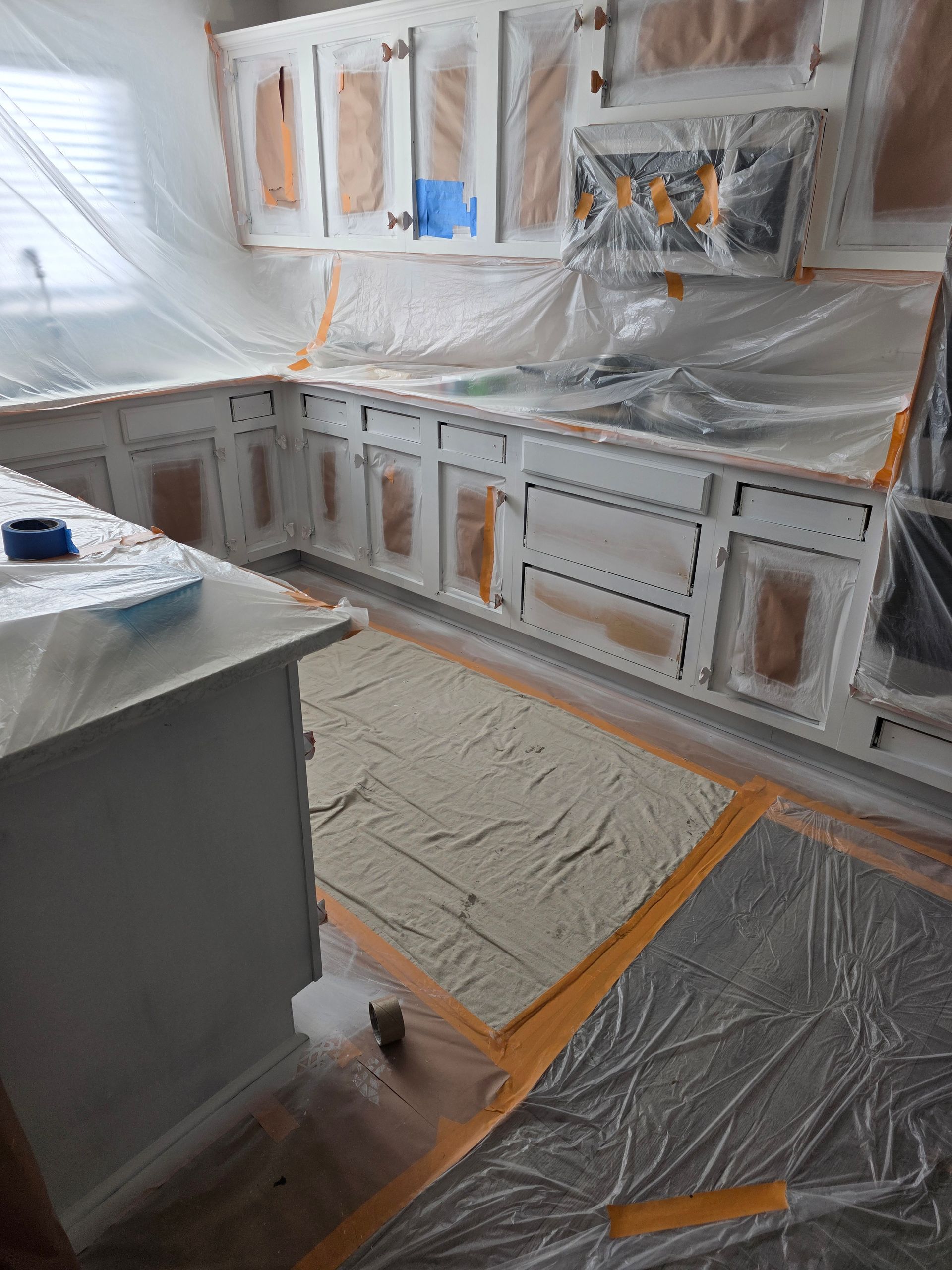 Kitchen cabinets being painted white, masked with plastic and tape to protect surrounding areas.