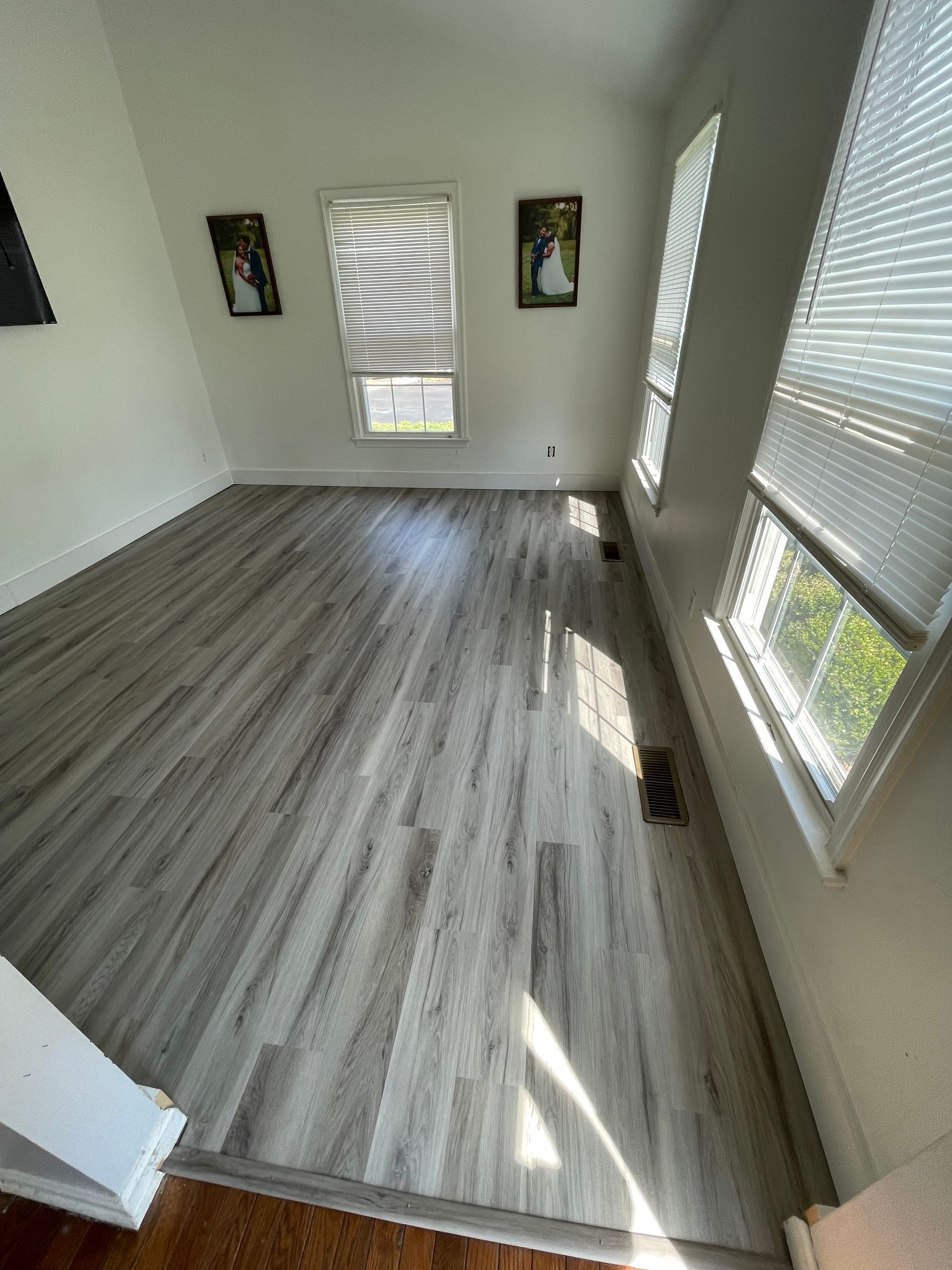 Empty room with gray wood-look flooring, two windows with blinds, and two framed pictures on a white wall.