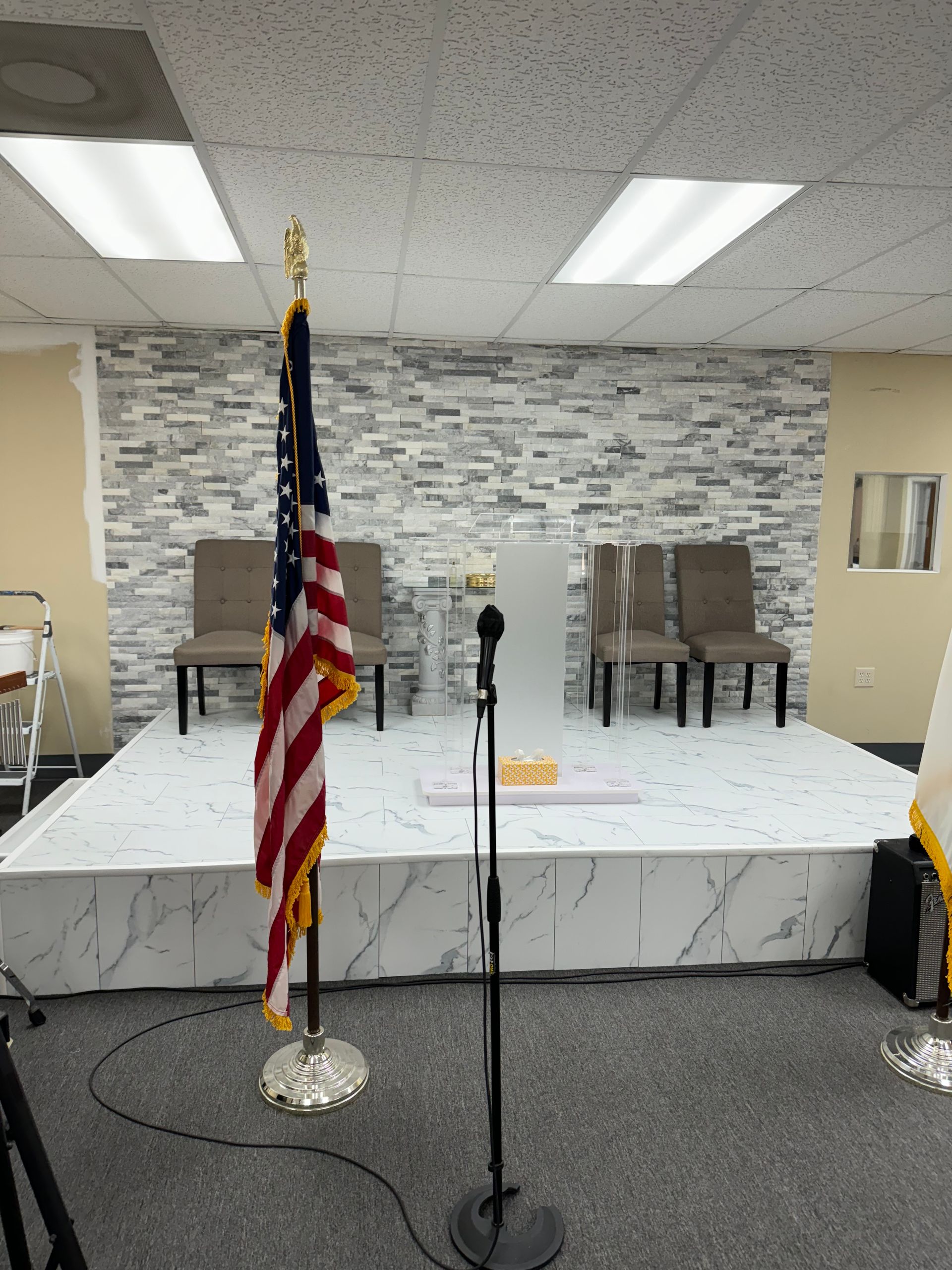 American flag on a stage with a microphone, chairs, and a white and gray tiled backdrop.