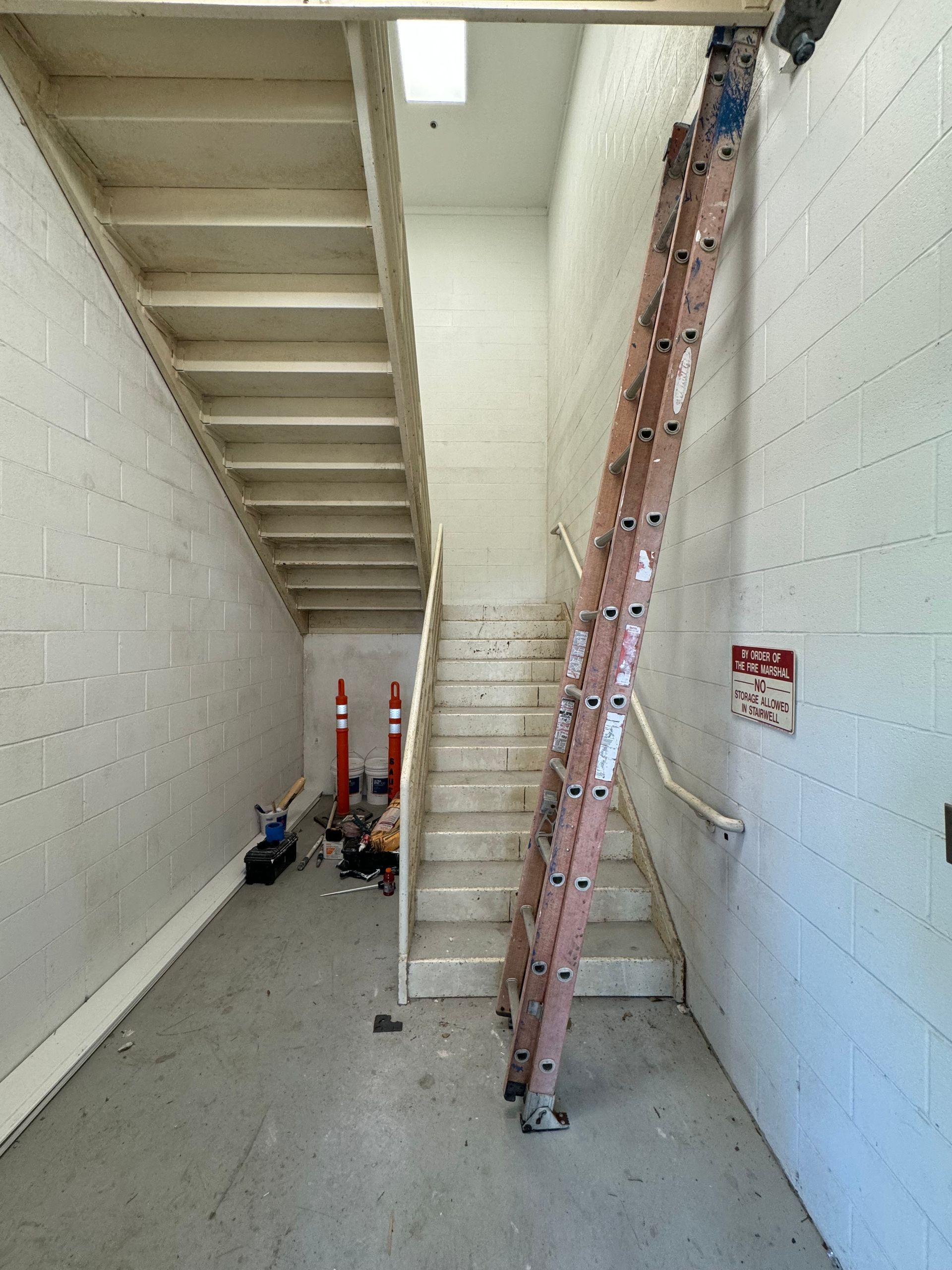 Ladder leaning against wall in stairwell; tools and debris on the floor, red and white safety sign.