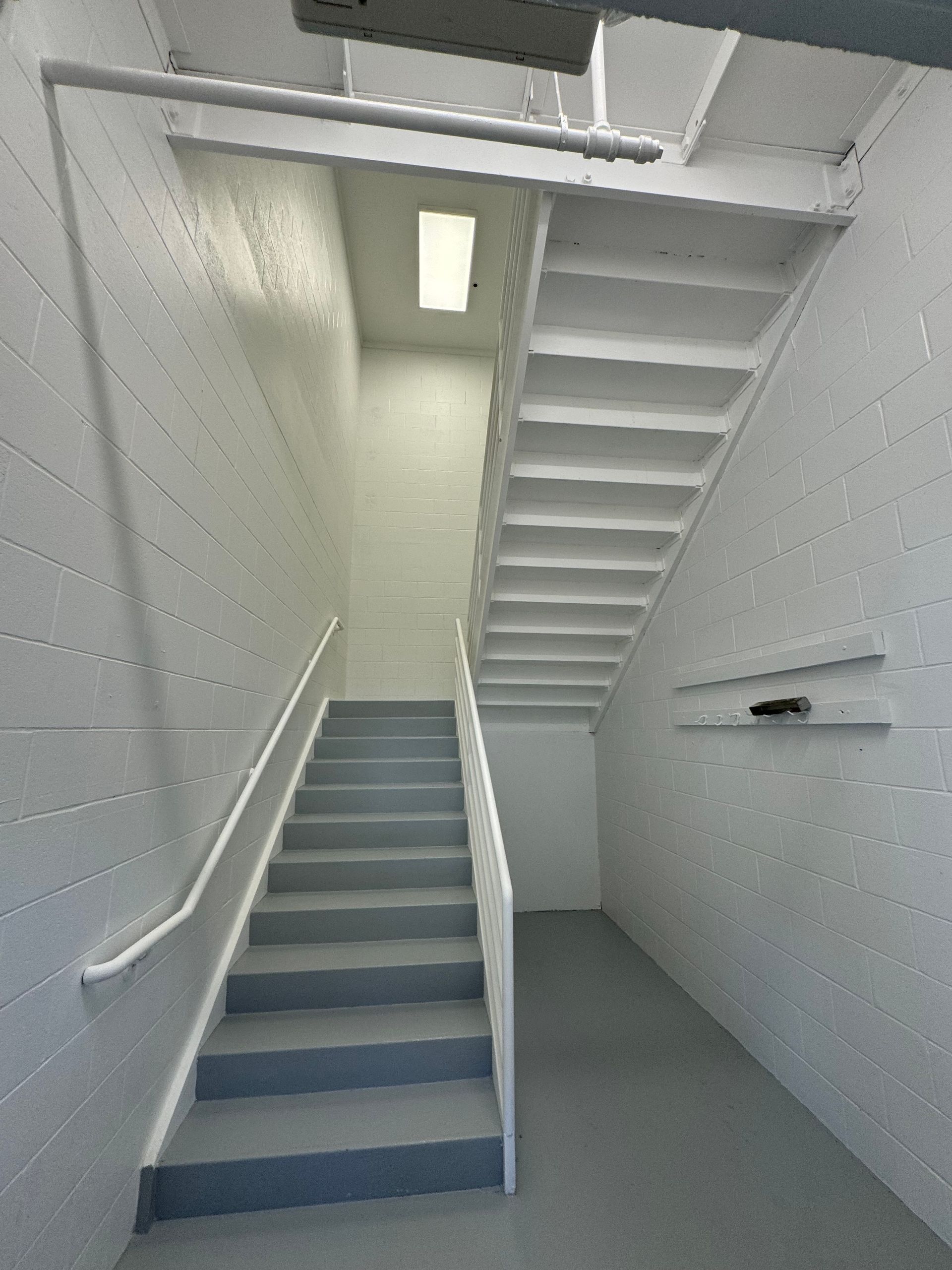 Staircase painted white and gray. Handrails are present on both sides. Overhead lighting in the stairwell.