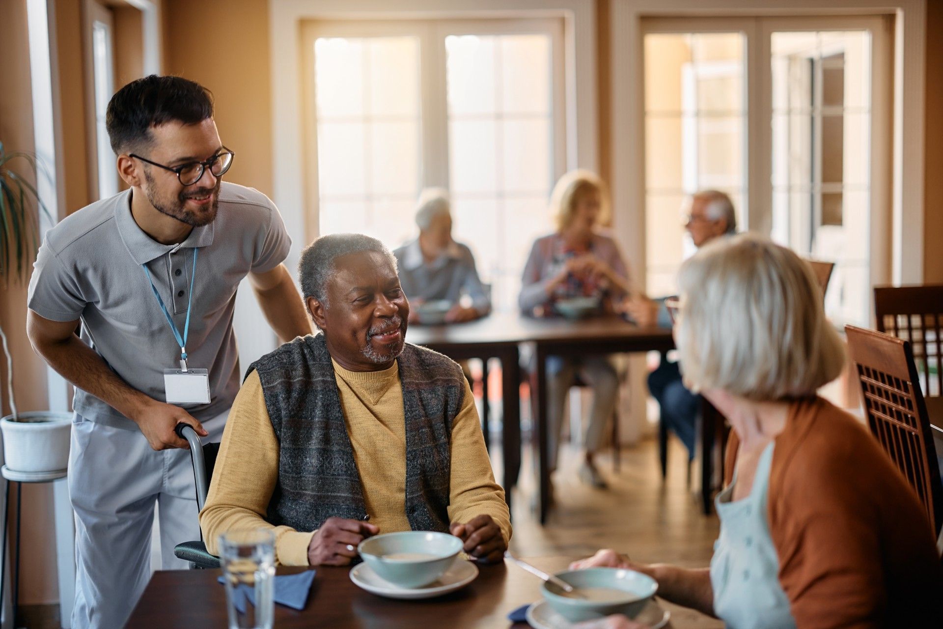 A man is talking to an elderly man sitting at a table in a nursing home.