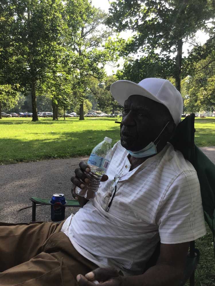 A man wearing a mask and a hat is sitting in a chair in a park.