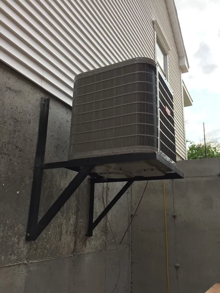 Air conditioning unit mounted on a black metal bracket attached to a concrete wall outside a building.