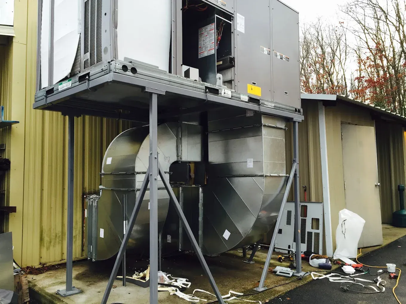 An industrial HVAC unit on a metal platform with ductwork, set against a yellow building and outdoor setting.