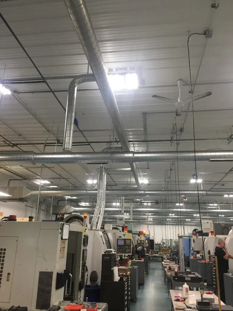 Factory interior with metal ductwork, machines, and overhead lighting.