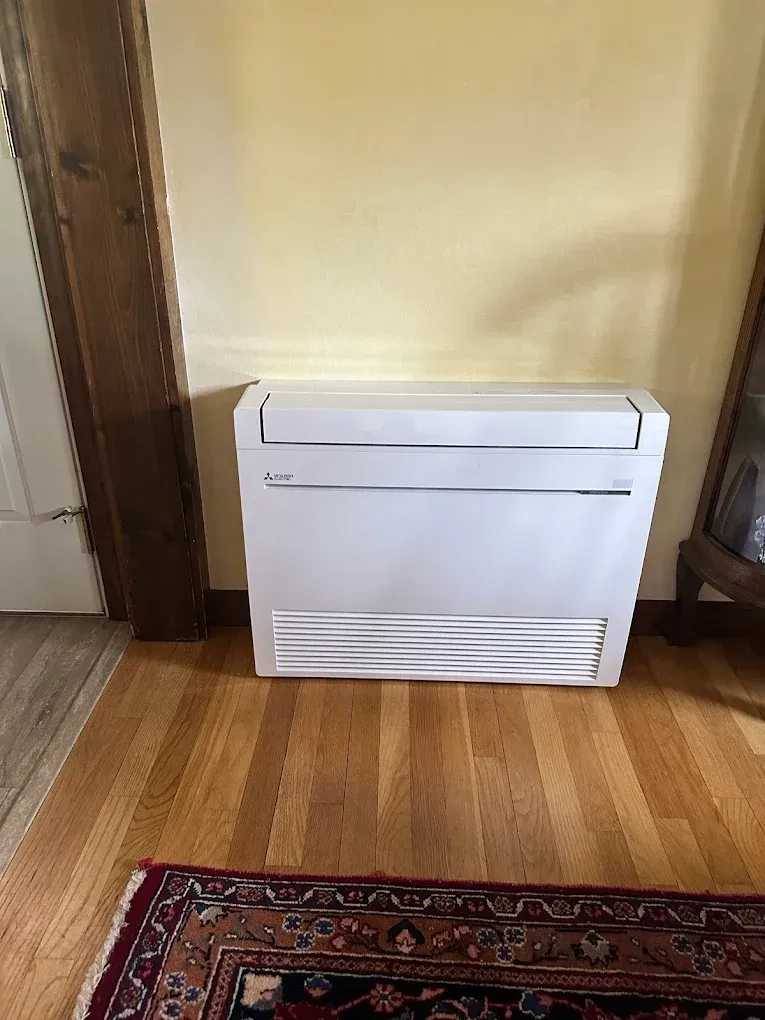 White air conditioning unit on wood floor, against a yellow wall, next to a wooden doorframe.