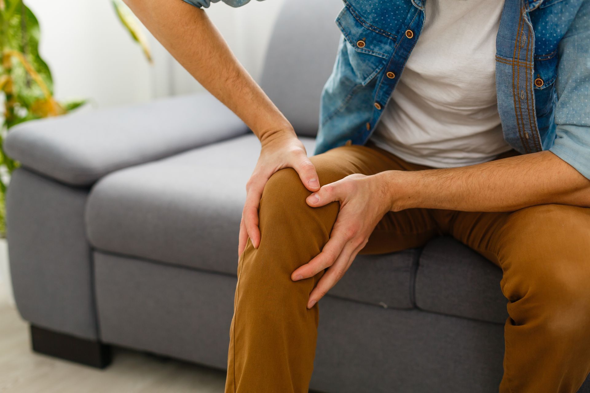 Man seated on a gray couch, holding his knee, likely experiencing pain. He wears a blue denim jacket, white shirt, and brown pants.