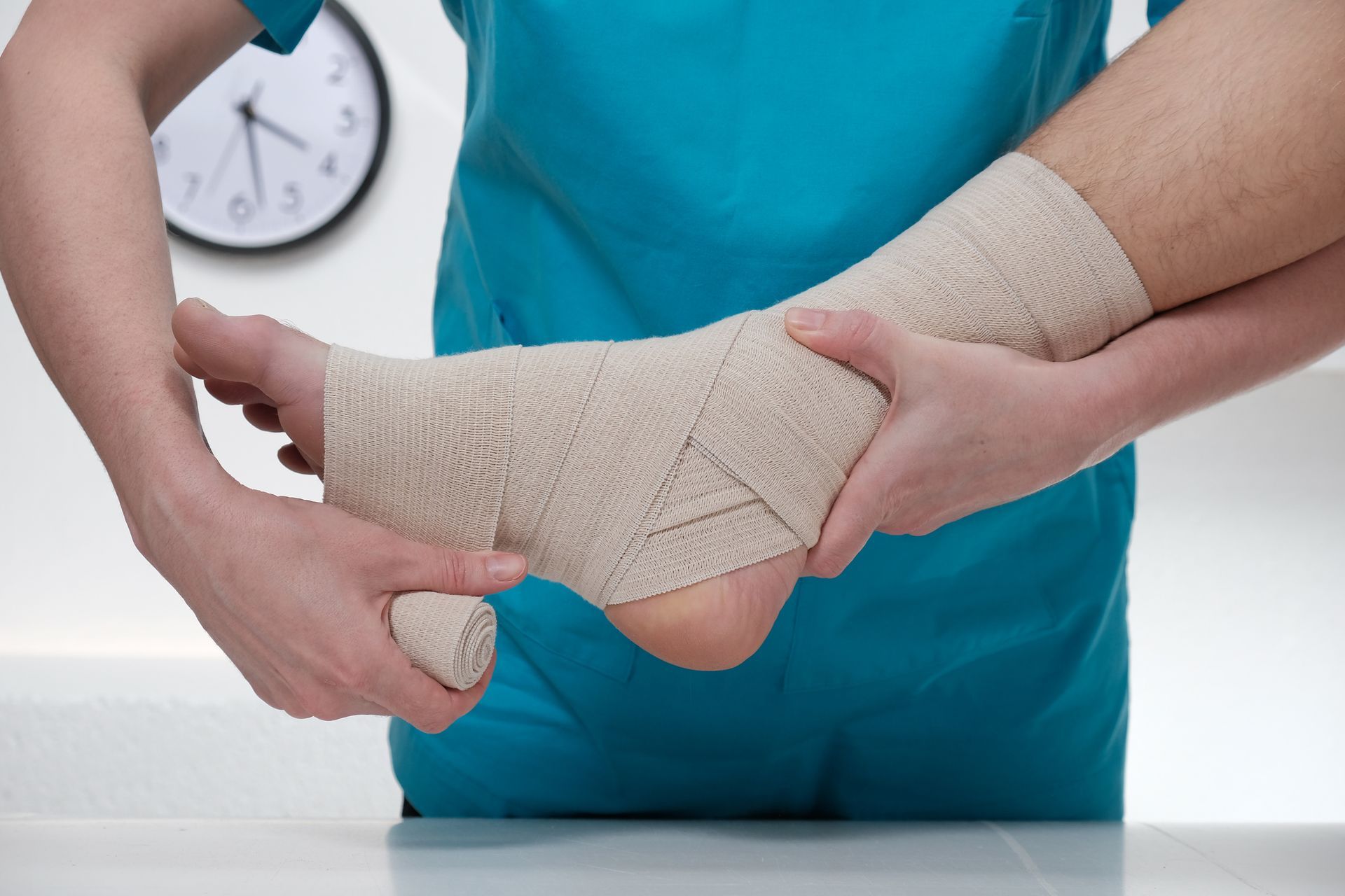 Person wrapping a beige bandage around someone's ankle and foot; a clock hangs on the wall in the background.