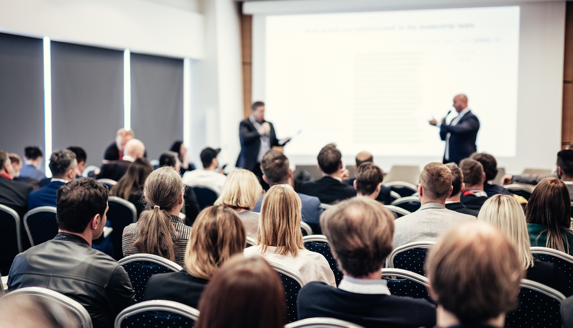 Audience seated, listening to two presenters on a stage with a screen in a conference room.