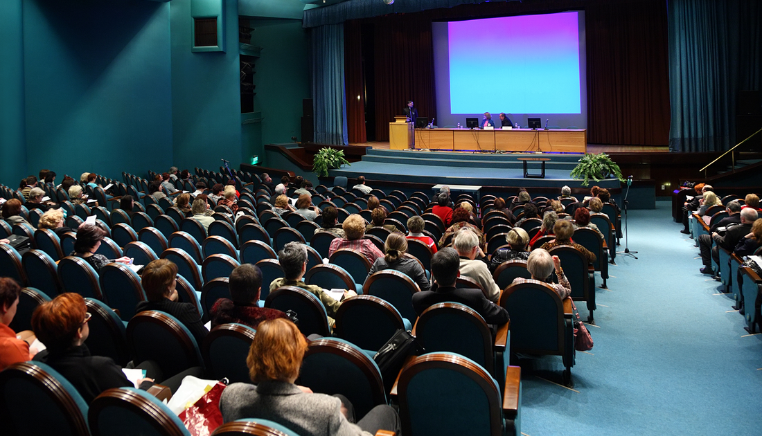 Audience seated in a large auditorium, watching a presentation on stage with a screen displaying blue hues.