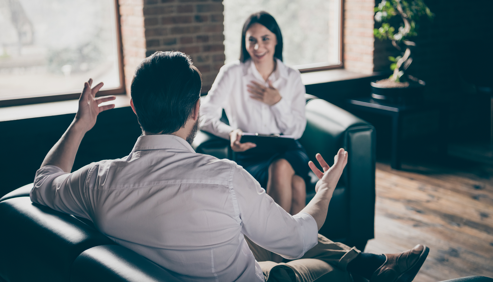 Man talking, gesturing with hands to smiling therapist holding a clipboard. In an office setting.