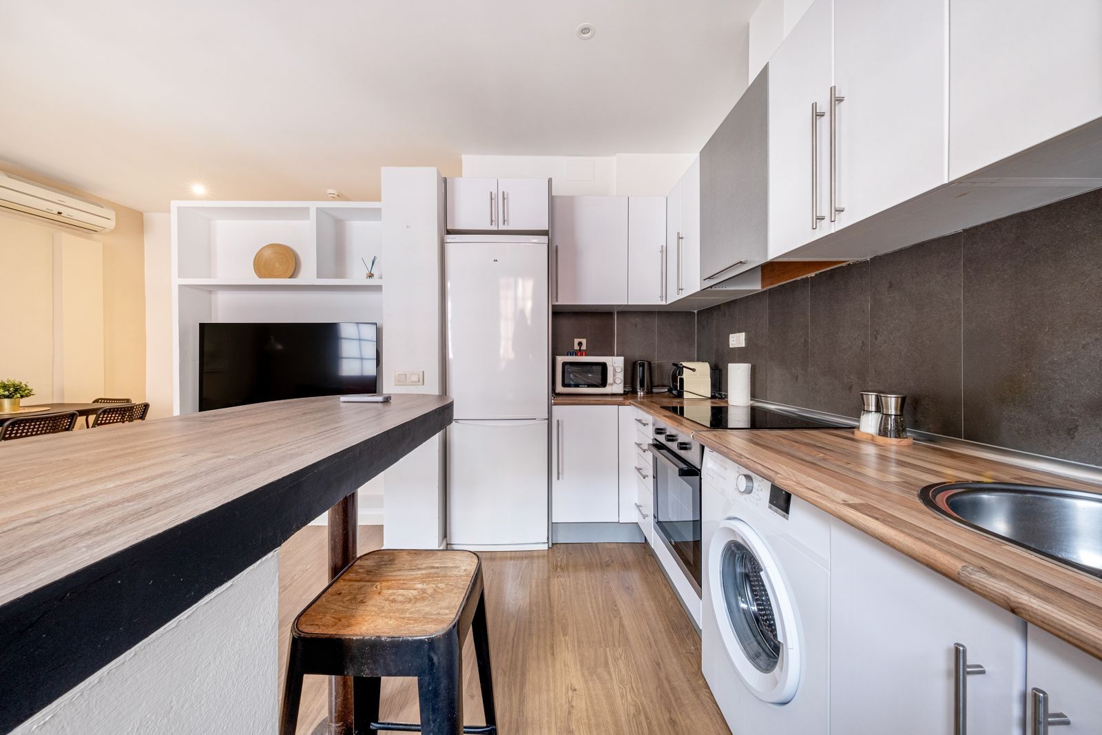 A kitchen with white cabinets , a washing machine , a refrigerator , a microwave and a television.