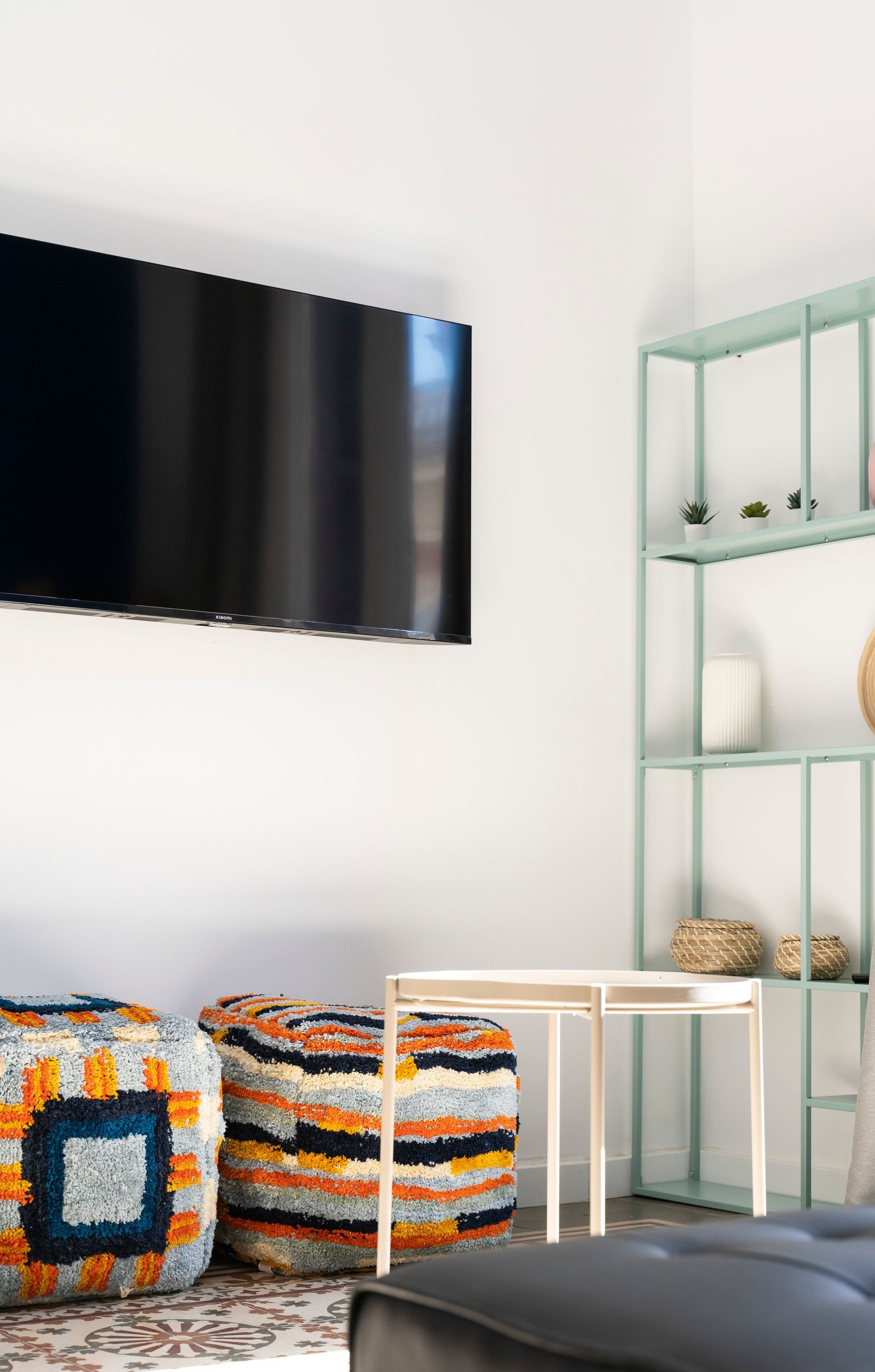 A living room with a flat screen tv , ottomans , a table and a shelf.