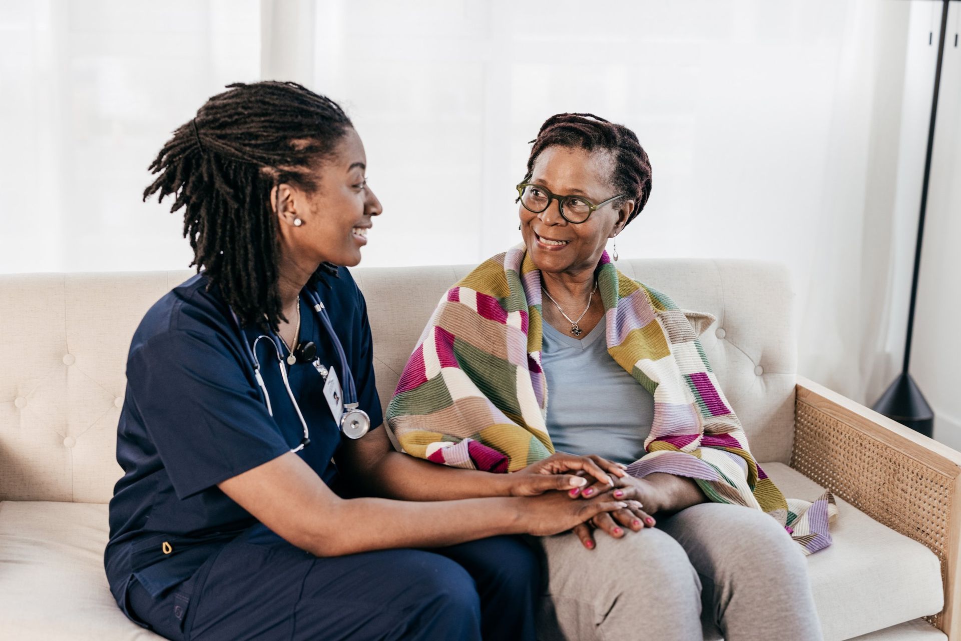 A nurse is holding the hand of an elderly woman while sitting on a couch.